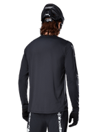 Alpinestars A-Dura Switch Langarmtrikot, Schwarz, Rückansicht eines Radfahrers mit schwarzem MTB-Helm und Handschuhen, mit technischem Jacquard-Gewebe und verlängertem Rückenteil für Offroad-Fahrten