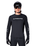 Alpinestars A-Dura Switch Langarmtrikot, schwarzes MTB-Trikot, weißer Alpinestars-Schriftzug auf der Brust und Sterngrafiken auf den Ärmeln, getragen mit schwarzem Mountainbike-Helm und Performance-Sonnenbrille, technische Trail-Bekleidung