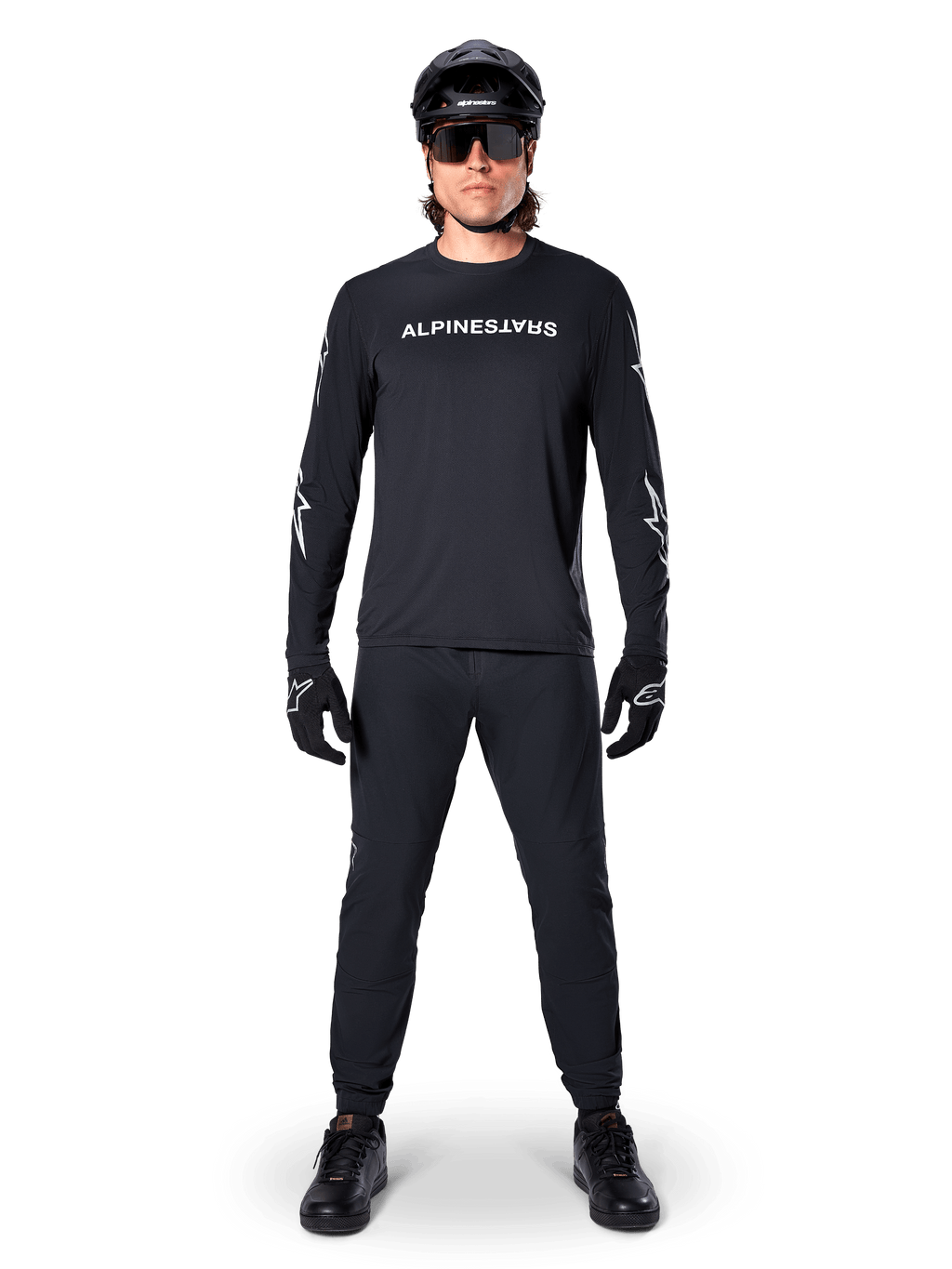 Alpinestars A-Dura Switch Langarmtrikot, Schwarz, getragen von einem Mountainbike-Fahrer mit passender schwarzer Hose und Helm, mit weißem Wortmarken-Logo auf der Brust, technische Offroad-Radsportbekleidung