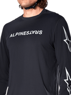 Alpinestars A-Dura Switch Langarmtrikot, Schwarz, mit weißem Alpinestars-Schriftzug auf der Brust und Sternenlogos auf dem Ärmel, getragen von einem Fahrer mit sichtbarem Helmriemen, funktionelle Mountainbike-Bekleidung