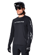 Männlicher Radfahrer im Alpinestars A-Dura Switch Langarmtrikot, Schwarz mit weißem Schriftzug und Stern-Logos auf den Ärmeln, kombiniert mit schwarzem MTB-Helm und Sonnenbrille, Hochleistungs-Radsportausrüstung für Offroad-Fahrten