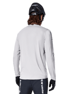 Alpinestars A-Dura Switch Langarmtrikot, Hellgrau, Rückansicht eines Mountainbikers mit schwarzem MTB-Helm und Handschuhen, mit verlängertem Rückensaum für zusätzliche Abdeckung und strukturiertem Jacquard-Gewebe, Performance-Radsportbekleidung.