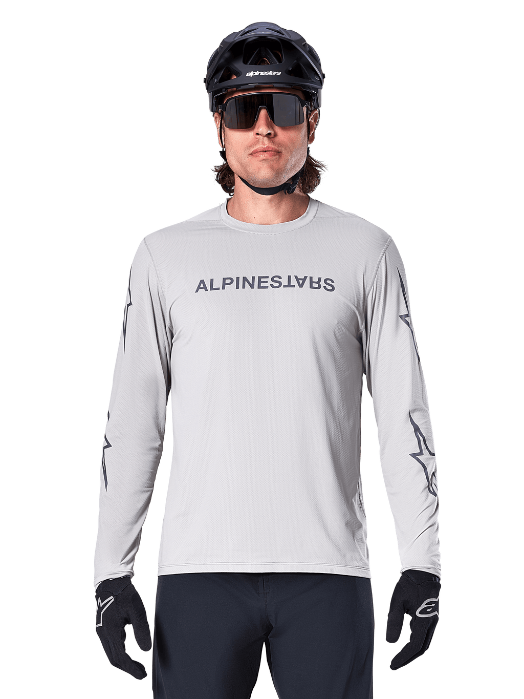 Alpinestars A-Dura Switch Langarmtrikot, Hellgrau, mit dunkelgrauem Schriftzug auf der Brust und Stern-Logos an den Ärmeln, getragen von einem Fahrer mit schwarzem MTB-Helm und Handschuhen, Performance-Radsportbekleidung für Trailfahrten