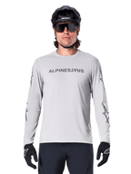 Alpinestars A-Dura Switch Langarmtrikot, Hellgrau, mit dunkelgrauem Schriftzug auf der Brust und Stern-Logos an den Ärmeln, getragen von einem Fahrer mit schwarzem MTB-Helm und Handschuhen, Performance-Radsportbekleidung für Trailfahrten