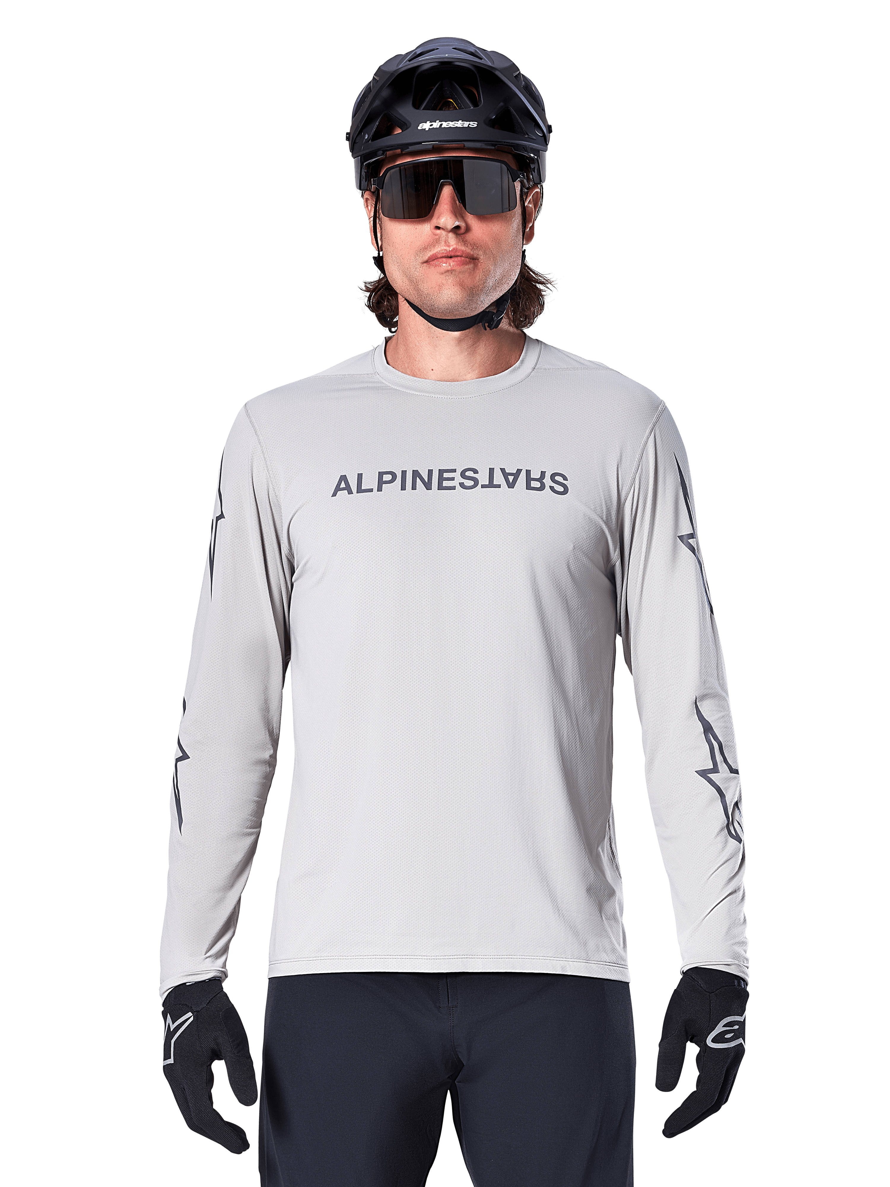 Alpinestars A-Dura Switch Langarmtrikot, Hellgrau, mit dunkelgrauem Schriftzug auf der Brust und Stern-Logos an den Ärmeln, getragen von einem Fahrer mit schwarzem MTB-Helm und Handschuhen, Performance-Radsportbekleidung für Trailfahrten