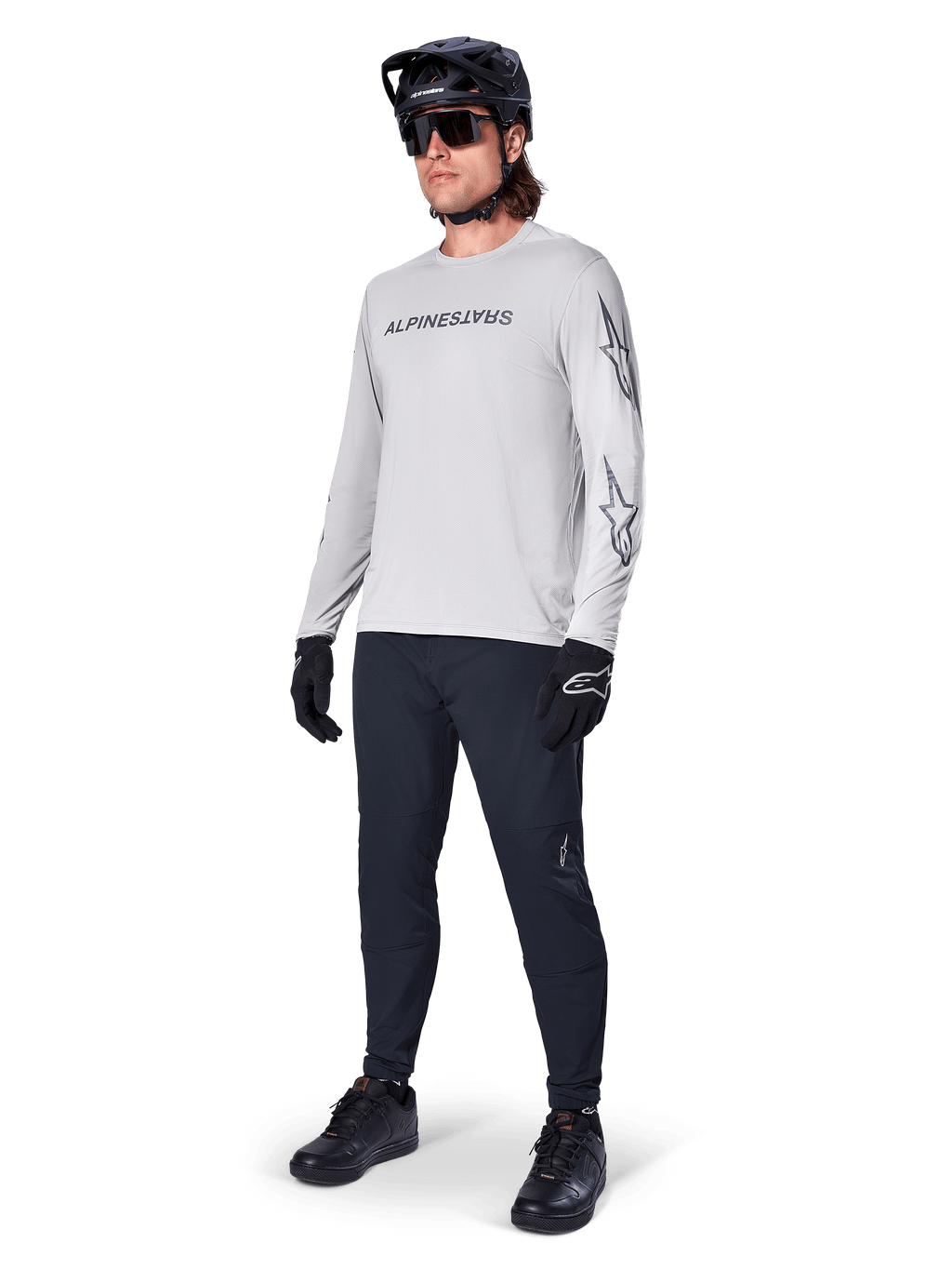 Alpinestars A-Dura Switch Langarmtrikot, Hellgrau, getragen von männlichem Fahrer mit schwarzer Mountainbike-Hose und Helm, schwarzes Wortmarken-Logo auf der Brust und Sterngrafiken auf den Ärmeln, Performance-Offroad-Radsportbekleidung