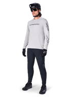 Alpinestars A-Dura Switch Langarmtrikot, Hellgrau, getragen von männlichem Fahrer mit schwarzer Mountainbike-Hose und Helm, schwarzes Wortmarken-Logo auf der Brust und Sterngrafiken auf den Ärmeln, Performance-Offroad-Radsportbekleidung