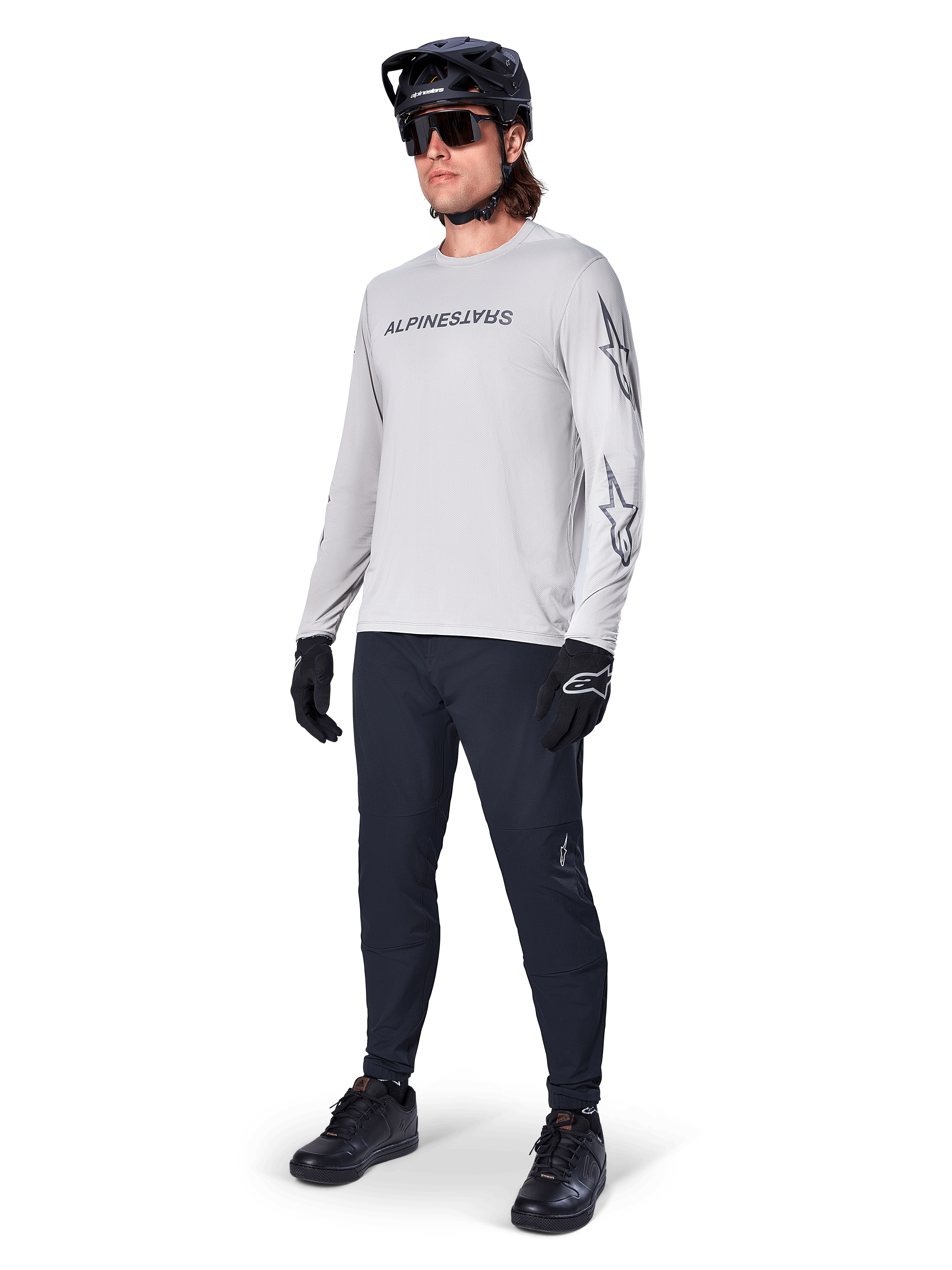 Alpinestars A-Dura Switch Langarmtrikot, Hellgrau, getragen von männlichem Fahrer mit schwarzer Mountainbike-Hose und Helm, schwarzes Wortmarken-Logo auf der Brust und Sterngrafiken auf den Ärmeln, Performance-Offroad-Radsportbekleidung