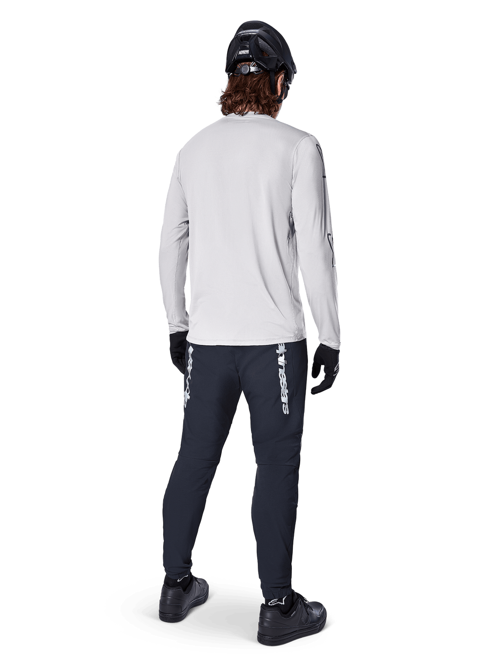 Alpinestars A-Dura Switch Langarmtrikot, Hellgrau, Rückansicht eines Fahrers mit schwarzer Radhose, Helm und Handschuhen, Hochleistungs-Mountainbike-Ausrüstung mit einem längeren abfallenden Saum.