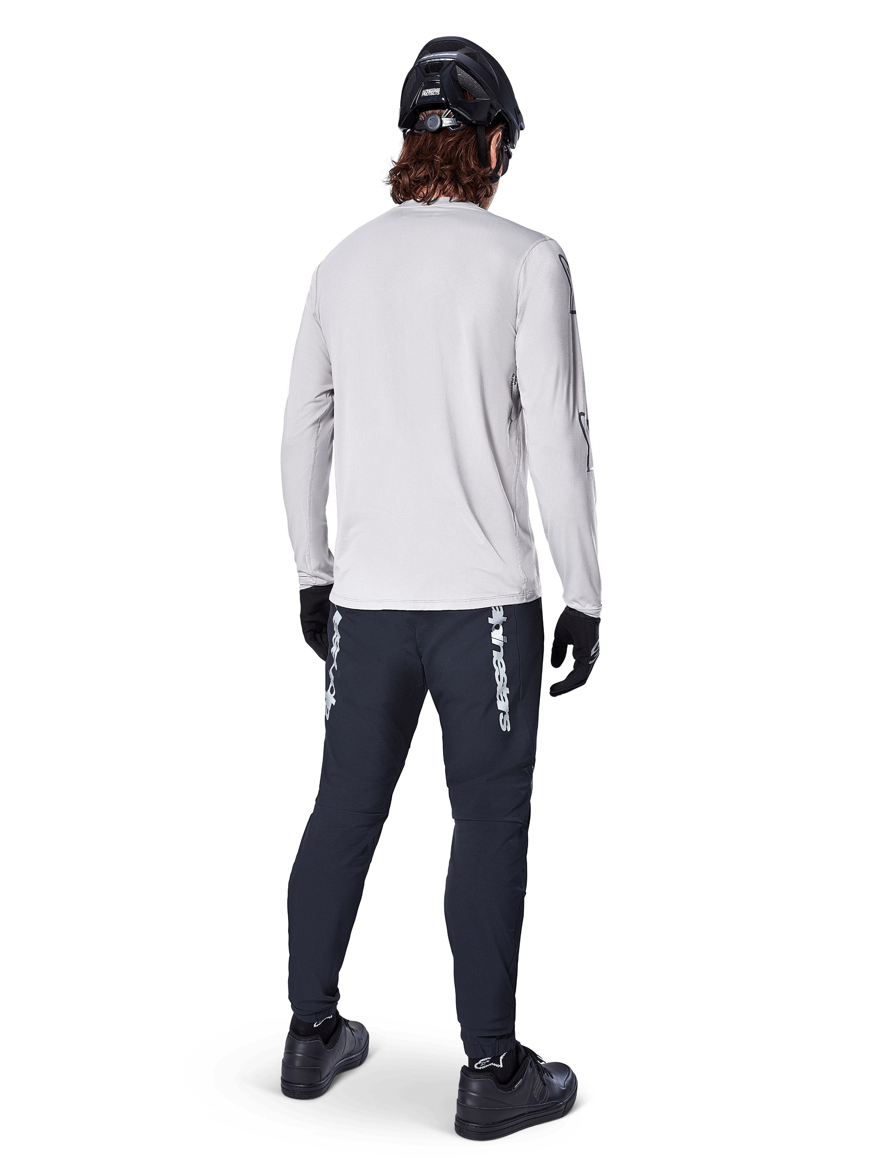 Alpinestars A-Dura Switch Langarmtrikot, Hellgrau, Rückansicht eines Fahrers mit schwarzer Radhose, Helm und Handschuhen, Hochleistungs-Mountainbike-Ausrüstung mit einem längeren abfallenden Saum.