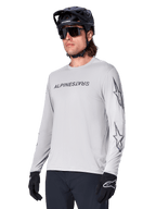 Alpinestars A-Dura Switch Langarmtrikot, Mountainbike-Trikot, Hellgrau, schwarzer Wortmarken-Siebdruck auf der Brust und Stern-Logos auf den Ärmeln, getragen mit schwarzem Mountainbike-Helm, dunkler Sonnenbrille und schwarzen Handschuhen, sportliche Offroad-Radfahrausrüstung