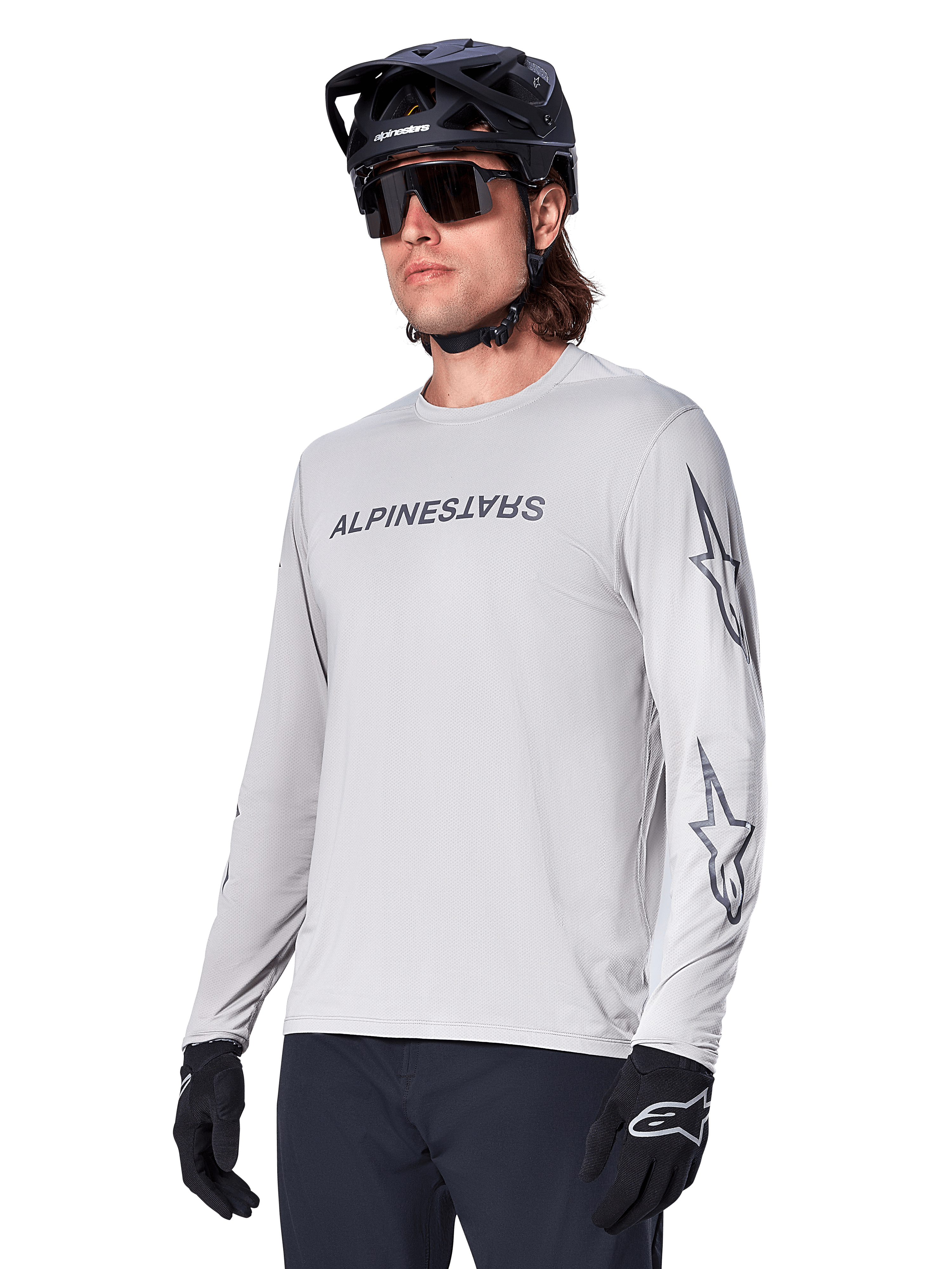 Alpinestars A-Dura Switch Langarmtrikot, Mountainbike-Trikot, Hellgrau, schwarzer Wortmarken-Siebdruck auf der Brust und Stern-Logos auf den Ärmeln, getragen mit schwarzem Mountainbike-Helm, dunkler Sonnenbrille und schwarzen Handschuhen, sportliche Offroad-Radfahrausrüstung