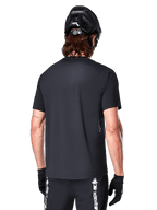 Alpinestars A-Dura Switch Kurzarmtrikot, Schwarz, Rückansicht eines Models mit schwarzem Fahrradhelm und MTB-Shorts, hebt die reguläre Passform und den verlängerten Rücken für das Mountainbiken hervor.