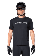 Mountainbike-Fahrer im Alpinestars A-Dura Switch Kurzarmtrikot in Schwarz, mit weißem Wortmarken-Logo auf der Brust, kombiniert mit schwarzem Helm, großer Sonnenbrille und schwarzen Handschuhen, professionelle Radsportbekleidung für Trail-Fahrten.