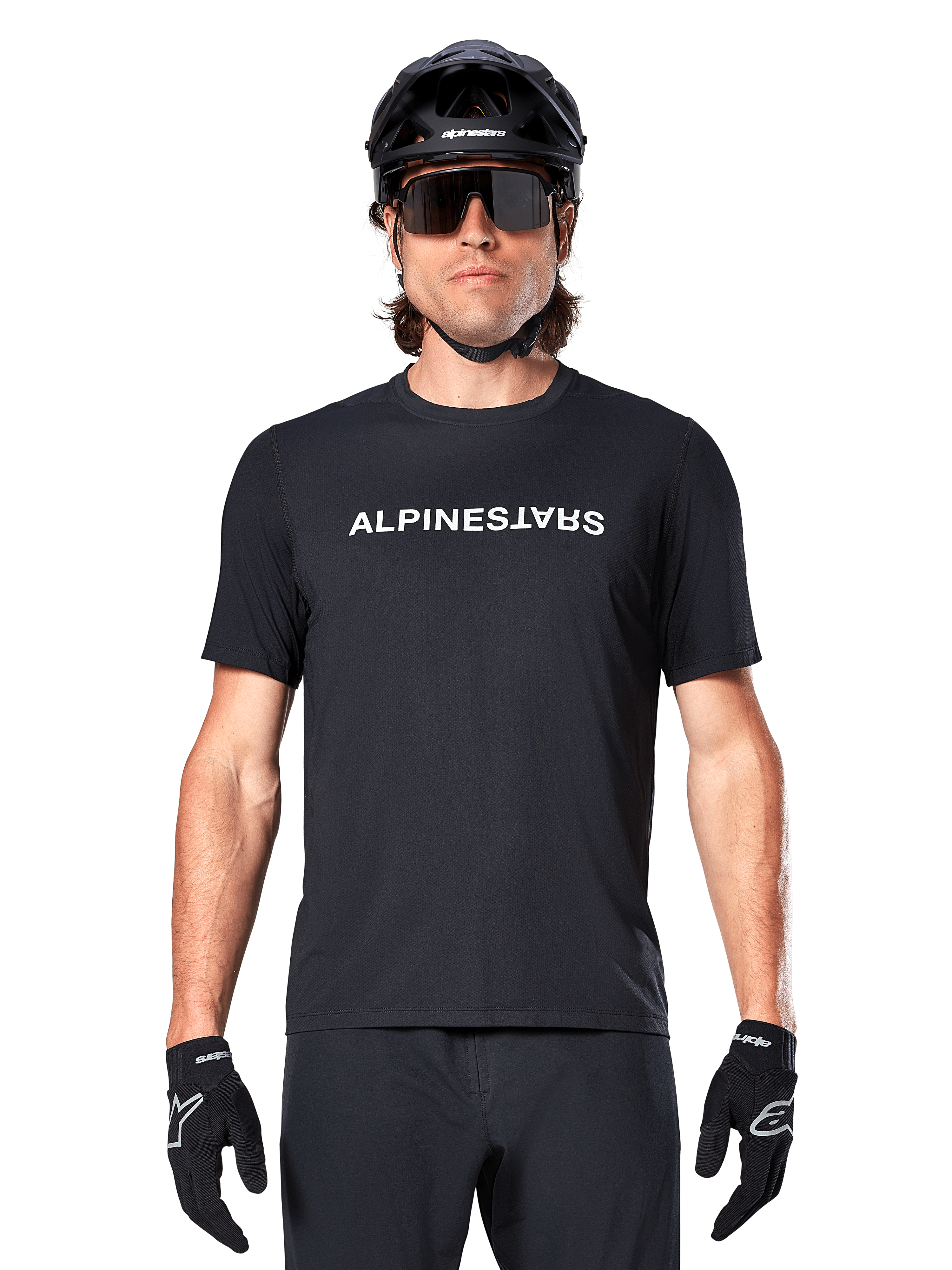Mountainbike-Fahrer im Alpinestars A-Dura Switch Kurzarmtrikot in Schwarz, mit weißem Wortmarken-Logo auf der Brust, kombiniert mit schwarzem Helm, großer Sonnenbrille und schwarzen Handschuhen, professionelle Radsportbekleidung für Trail-Fahrten.