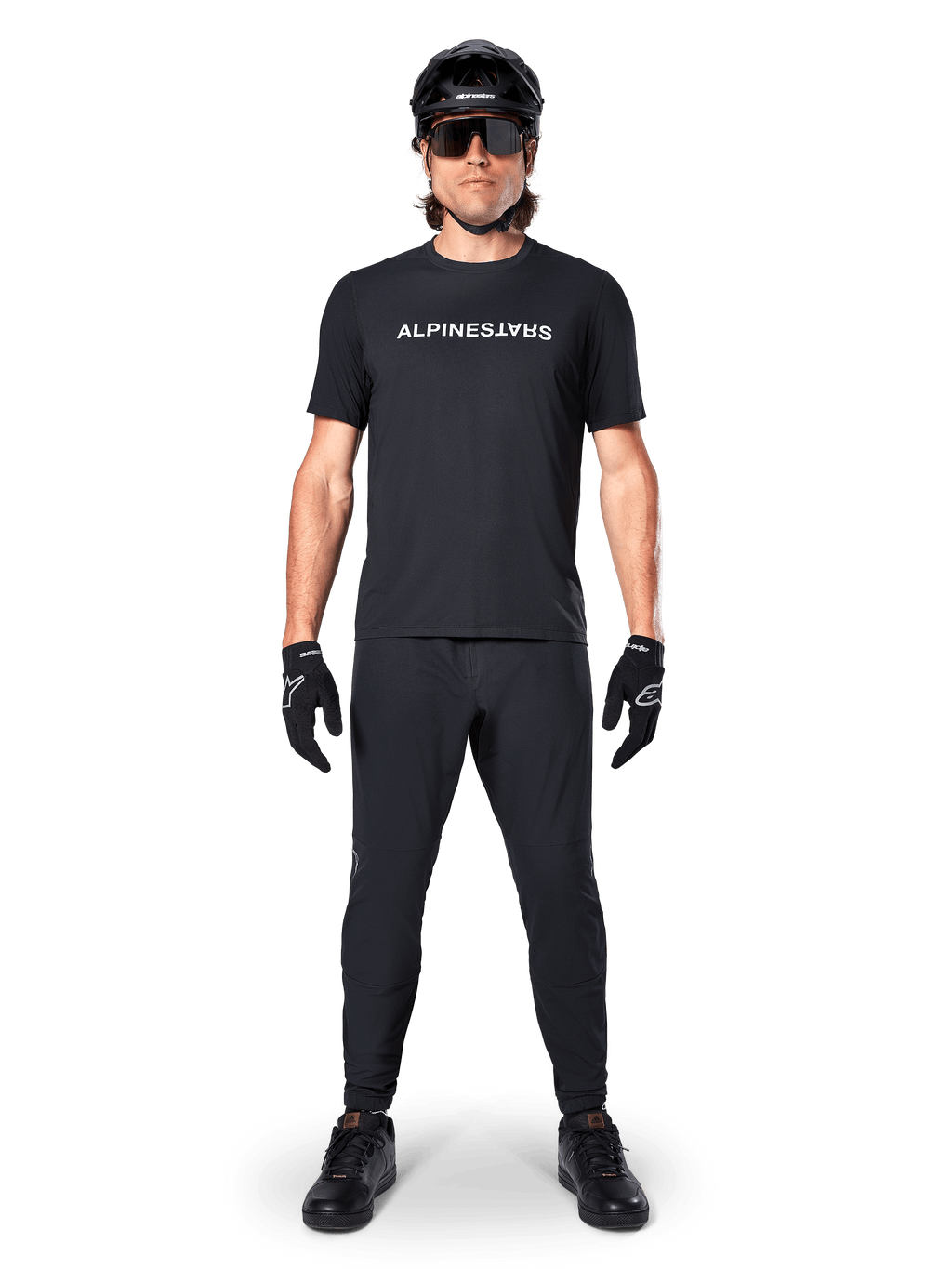 Mountainbike-Fahrer im schwarzen Alpinestars A-Dura Switch Kurzarmtrikot mit weißem Wortmarken-Logo auf der Brust, kombiniert mit passender schwarzer MTB-Hose, Helm und Handschuhen, komplettes trailbereites Radsport-Outfit.