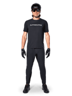 Mountainbike-Fahrer im schwarzen Alpinestars A-Dura Switch Kurzarmtrikot mit weißem Wortmarken-Logo auf der Brust, kombiniert mit passender schwarzer MTB-Hose, Helm und Handschuhen, komplettes trailbereites Radsport-Outfit.