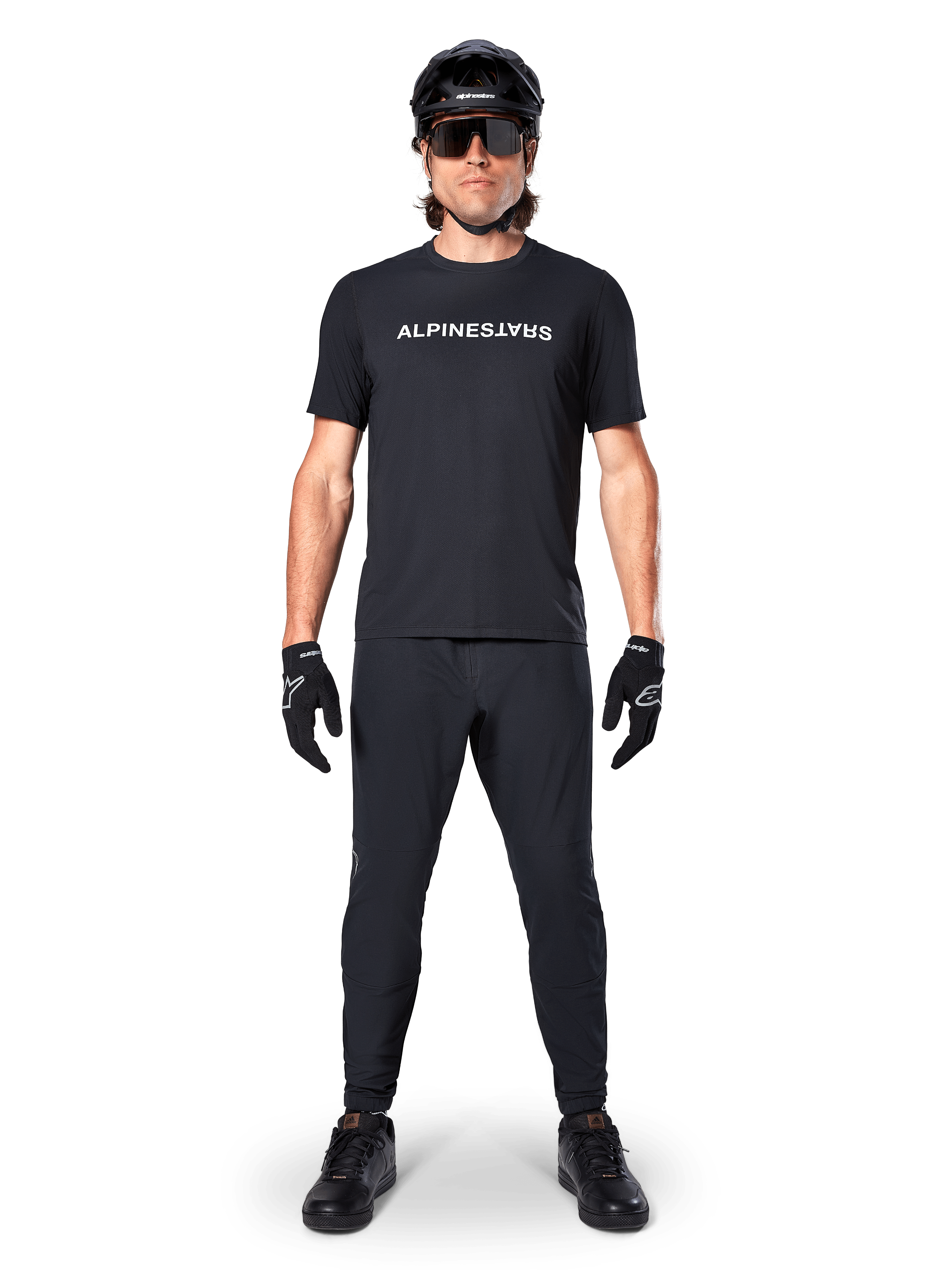 Mountainbike-Fahrer im schwarzen Alpinestars A-Dura Switch Kurzarmtrikot mit weißem Wortmarken-Logo auf der Brust, kombiniert mit passender schwarzer MTB-Hose, Helm und Handschuhen, komplettes trailbereites Radsport-Outfit.