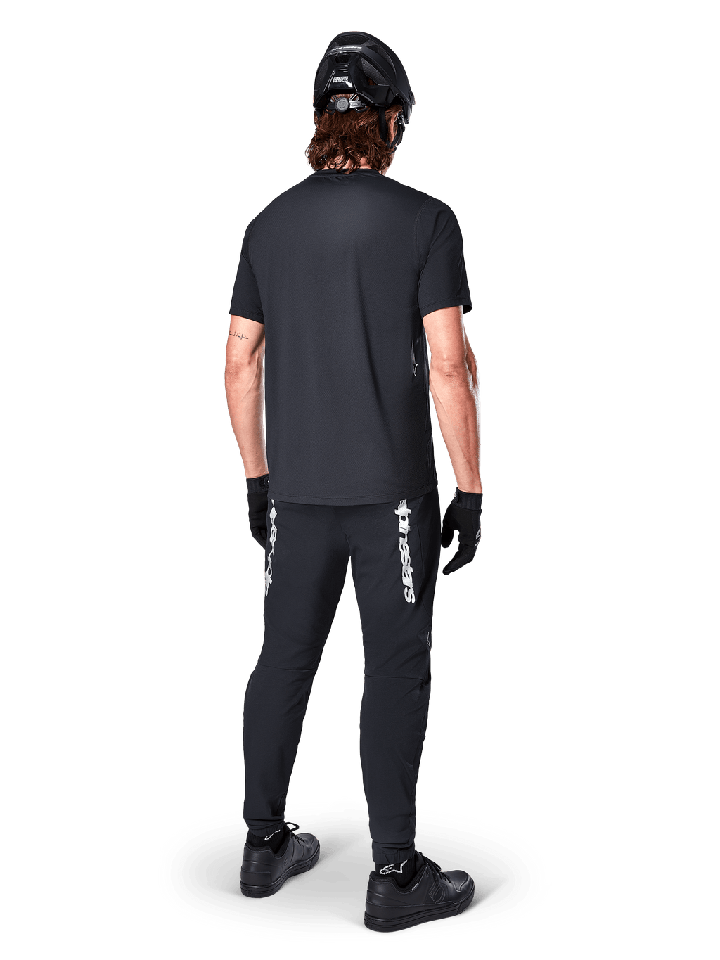 Alpinestars A-Dura Switch Kurzarmtrikot, Schwarz, Rückansicht eines Fahrers in kompletter Mountainbike-Ausrüstung, inklusive passender schwarzer Hose mit weißem Schriftzug, schwarzem Helm und Handschuhen, Performance-Radbekleidung für Trailfahrten