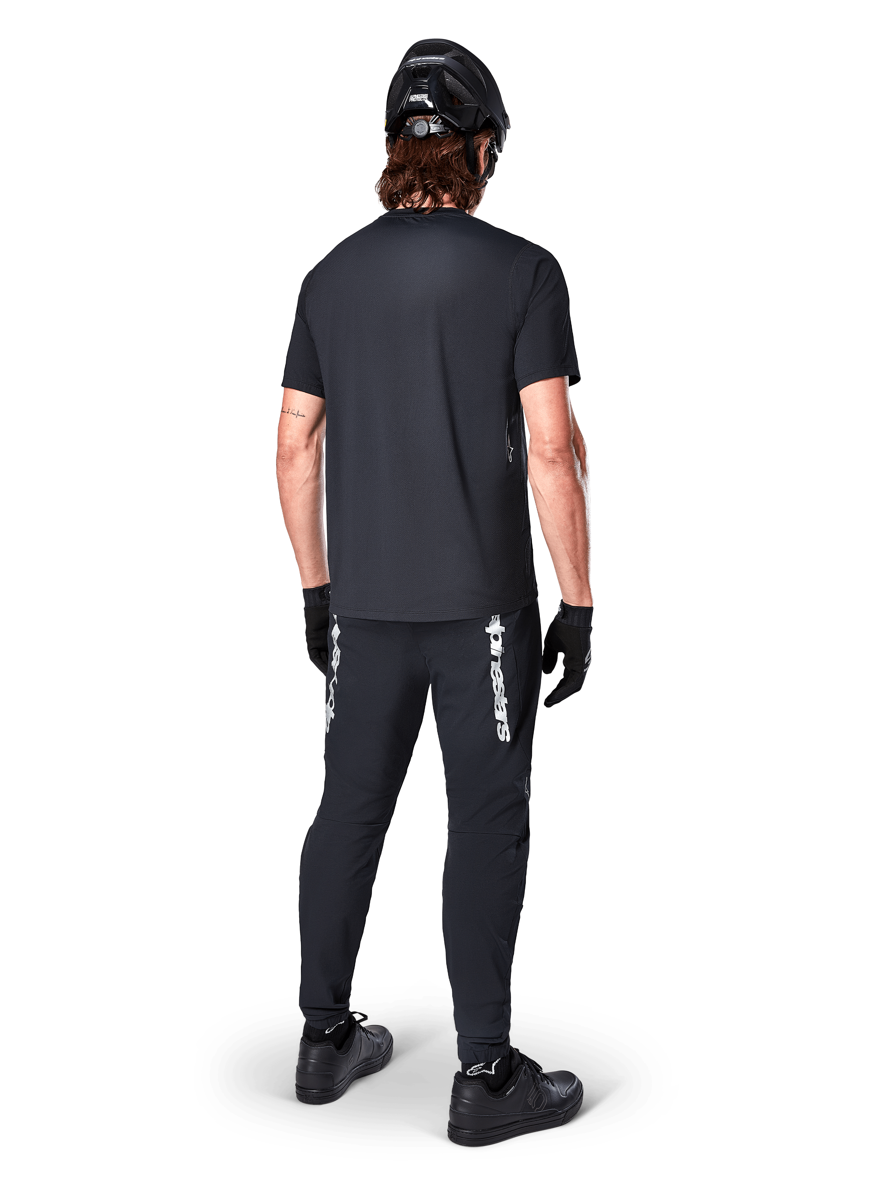 Alpinestars A-Dura Switch Kurzarmtrikot, Schwarz, Rückansicht eines Fahrers in kompletter Mountainbike-Ausrüstung, inklusive passender schwarzer Hose mit weißem Schriftzug, schwarzem Helm und Handschuhen, Performance-Radbekleidung für Trailfahrten