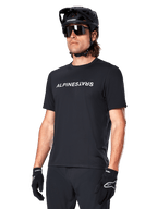 Alpinestars A-Dura Switch Kurzarmtrikot, Schwarz, mit weißem Wortmarken-Logo auf der Brust, getragen von einem Radfahrer mit schwarzem MTB-Helm und Sonnenbrille, technische Bekleidung für Trailriding