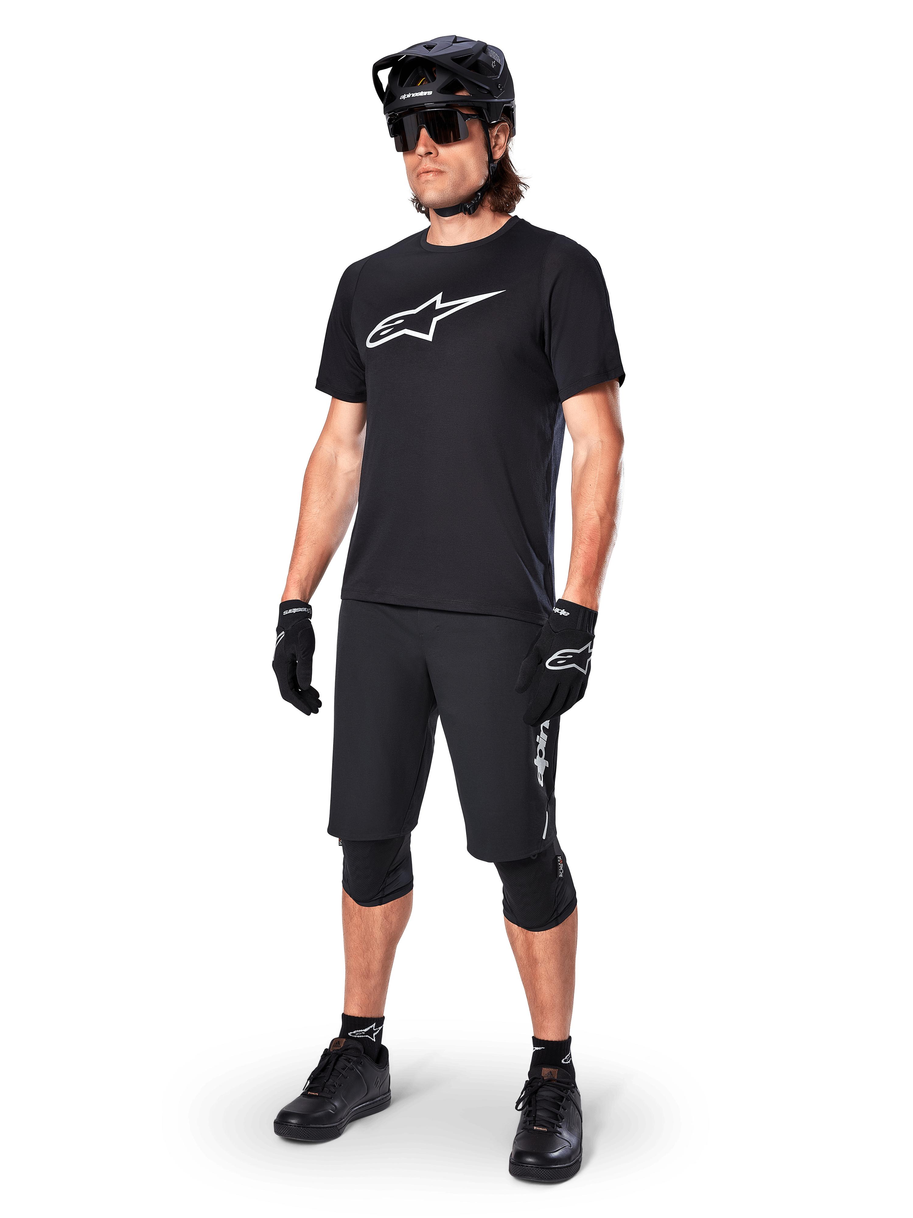 Mountain bike rider wearing the Alpinestars A-Dura Dri Oscar Short Sleeve Jersey in Black Reflective, featuring a large white logo on the chest, paired with matching black MTB shorts, gloves, and helmet, technical cycling apparel for off-road performance