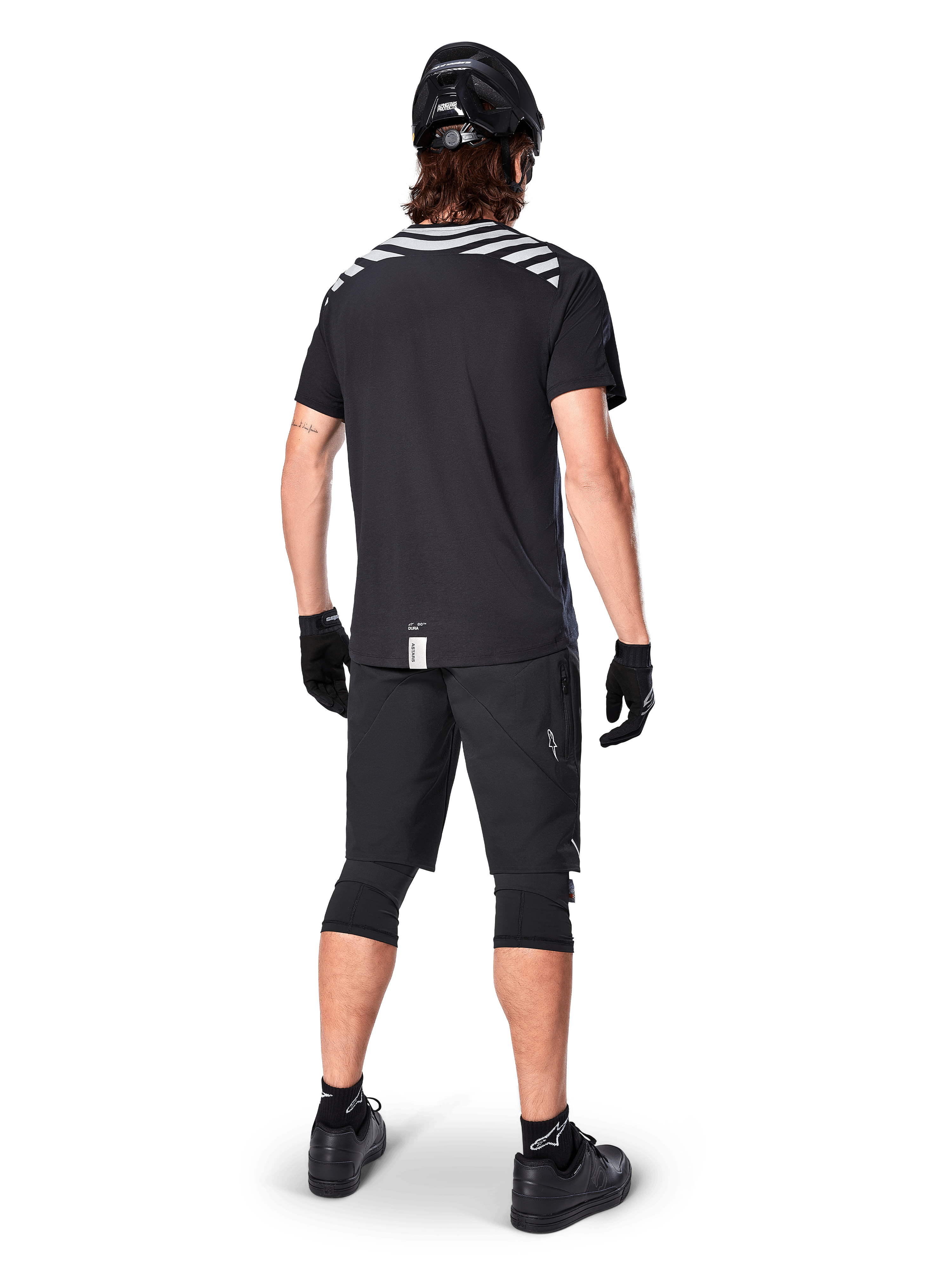 Alpinestars A-Dura Dri Oscar Short Sleeve Jersey, Black Reflective, rear view of a rider wearing the jersey with black mountain bike shorts, helmet, and knee pads, featuring reflective shoulder graphics and a drop tail design for MTB riding.