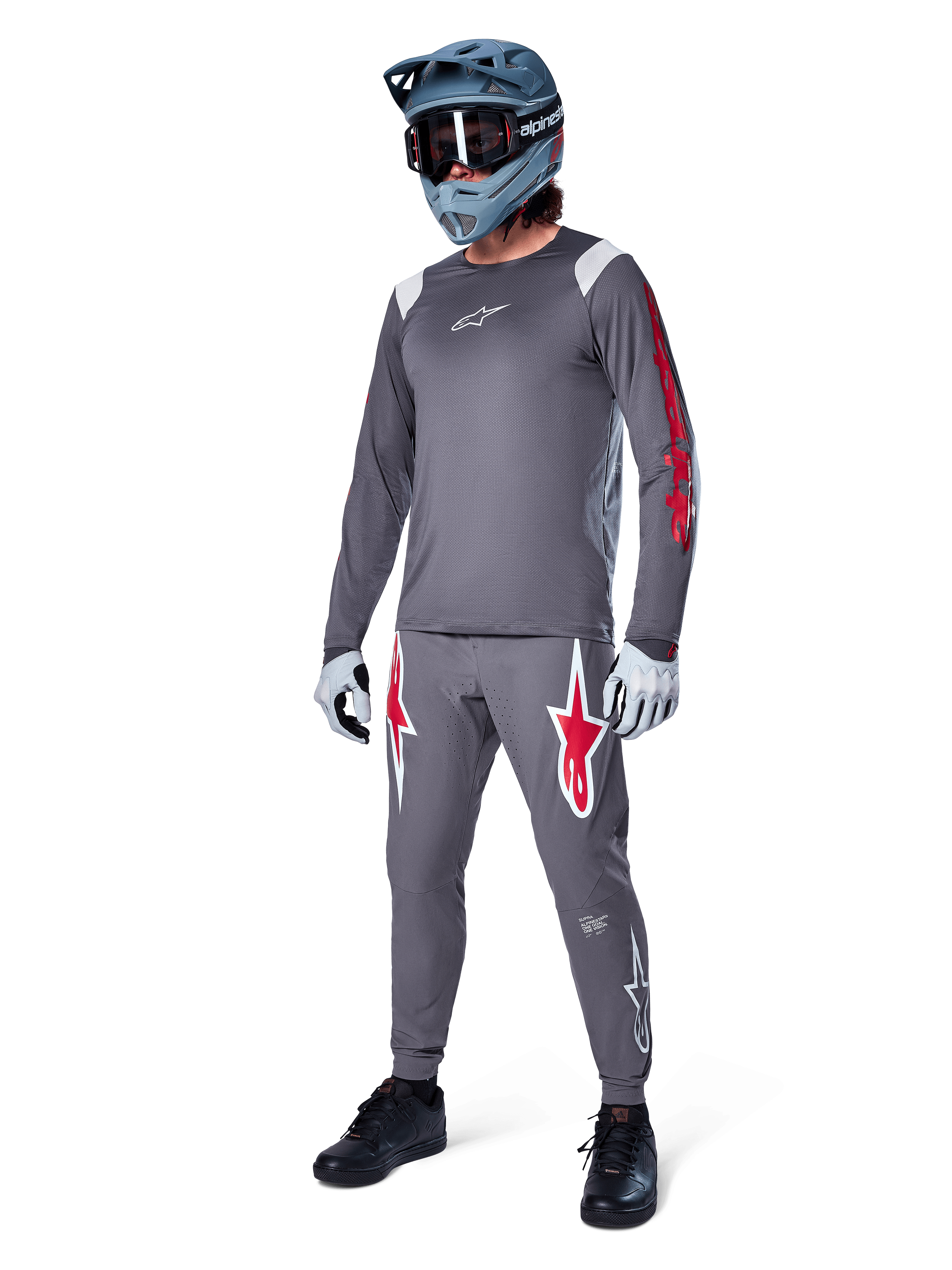 Alpinestars A-Supra Astar Langarmtrikot in Dunkelgrau, mit rotem Branding auf den Ärmeln und weißem Logo auf der Brust, getragen von einem Fahrer mit passender grauer Hose, einem blaugrauen Full-Face-Helm und Performance-Handschuhen, koordinierte Mountainbike-Ausrüstung für Gravity-Riding.