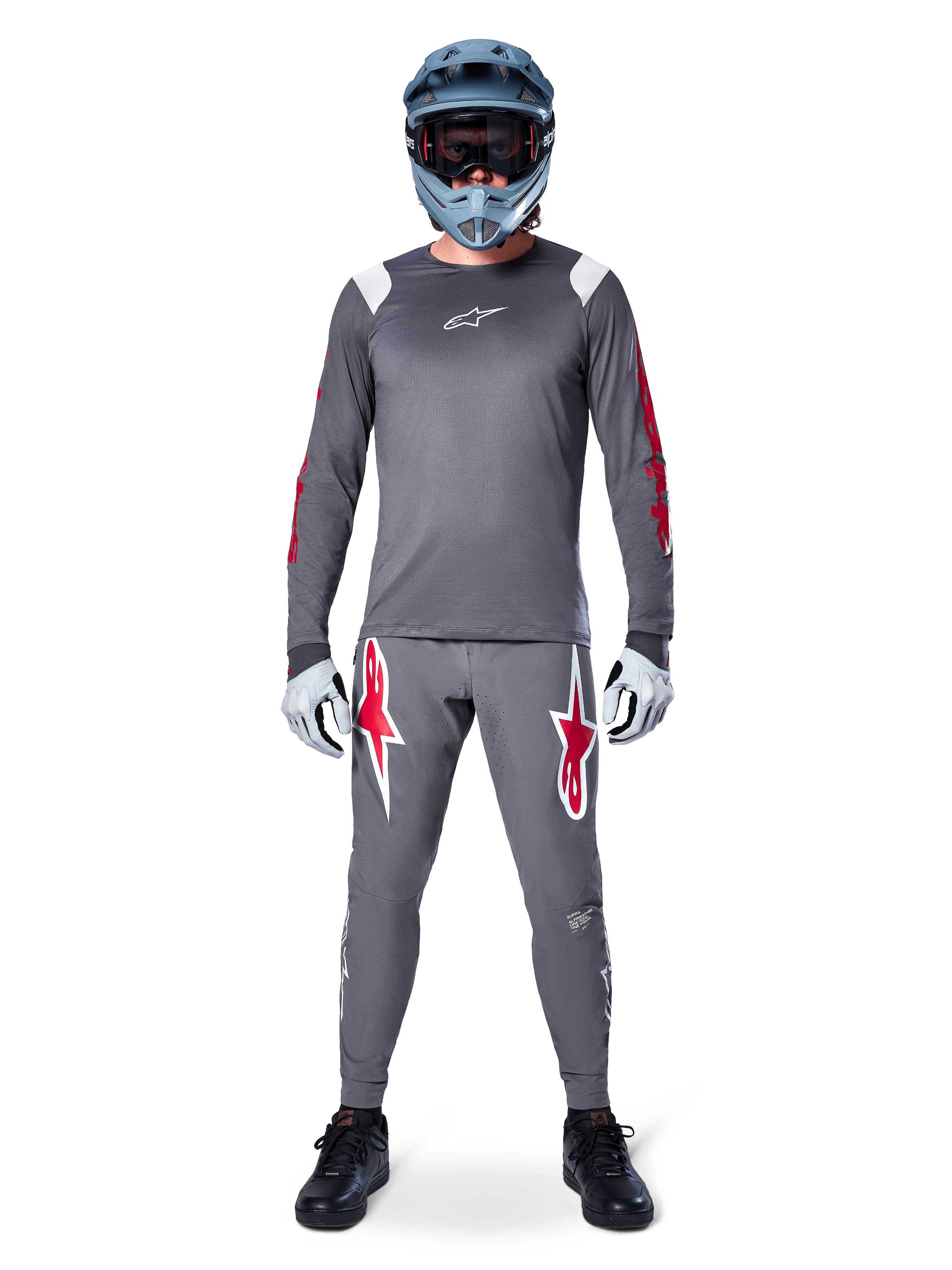 MTB-Fahrer im Alpinestars A-Supra Astar Langarmtrikot, Dunkelgrau mit roten Akzenten und weißem Brust-Logo, dazu passende graue Hose mit großen roten Sterngrafiken, blauer Fullface-Helm mit Goggle, hellgraue Handschuhe und schwarze Schuhe, professionelle Mountainbike-Ausrüstung