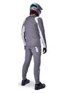 Alpinestars A-Supra Astar Trikot, Langarm-MTB-Trikot, Dunkelgrau mit weißen Ärmelakzenten und rotem Logo, Rückansicht eines Fahrers in passender grauer Hose und blauem Full-Face-Helm, Performance-Radsportbekleidung