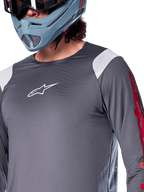 Alpinestars A-Supra Astar Langarmtrikot, Dunkelgrau mit weißen Schultereinsätzen und roten Grafiken am Ärmel, getragen mit einem hellblau-grauen Full-Face-Helm, Performance-Mountainbike-Bekleidung
