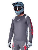Alpinestars A-Supra Astar Langarmtrikot, MTB-Trikot, Dunkelgrau mit roten und weißen Akzenten, Performance-Passform, getragen mit passender grauer Hose mit roten und weißen Sterngrafiken, komplettes Offroad-Cycling-Outfit mit Full-Face-Helm und Handschuhen