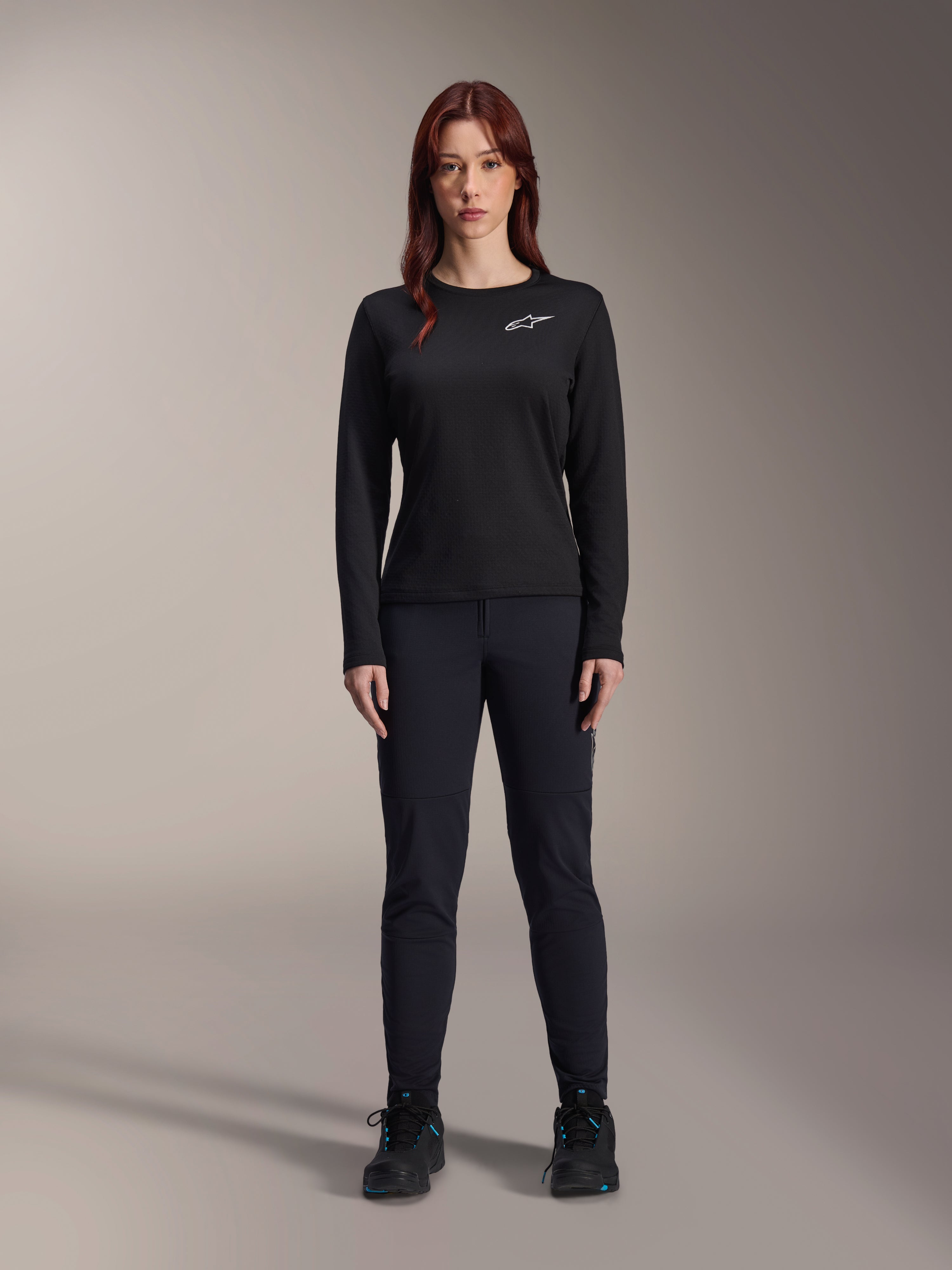 Alpinestars Stella A-Dura Thermo-Trikot, langärmeliges Damen-Mountainbike-Trikot in Schwarz, weißes Logo auf der Brust, kombiniert mit passender schwarzer Ausrüstung und Radschuhen, Studioporträt.