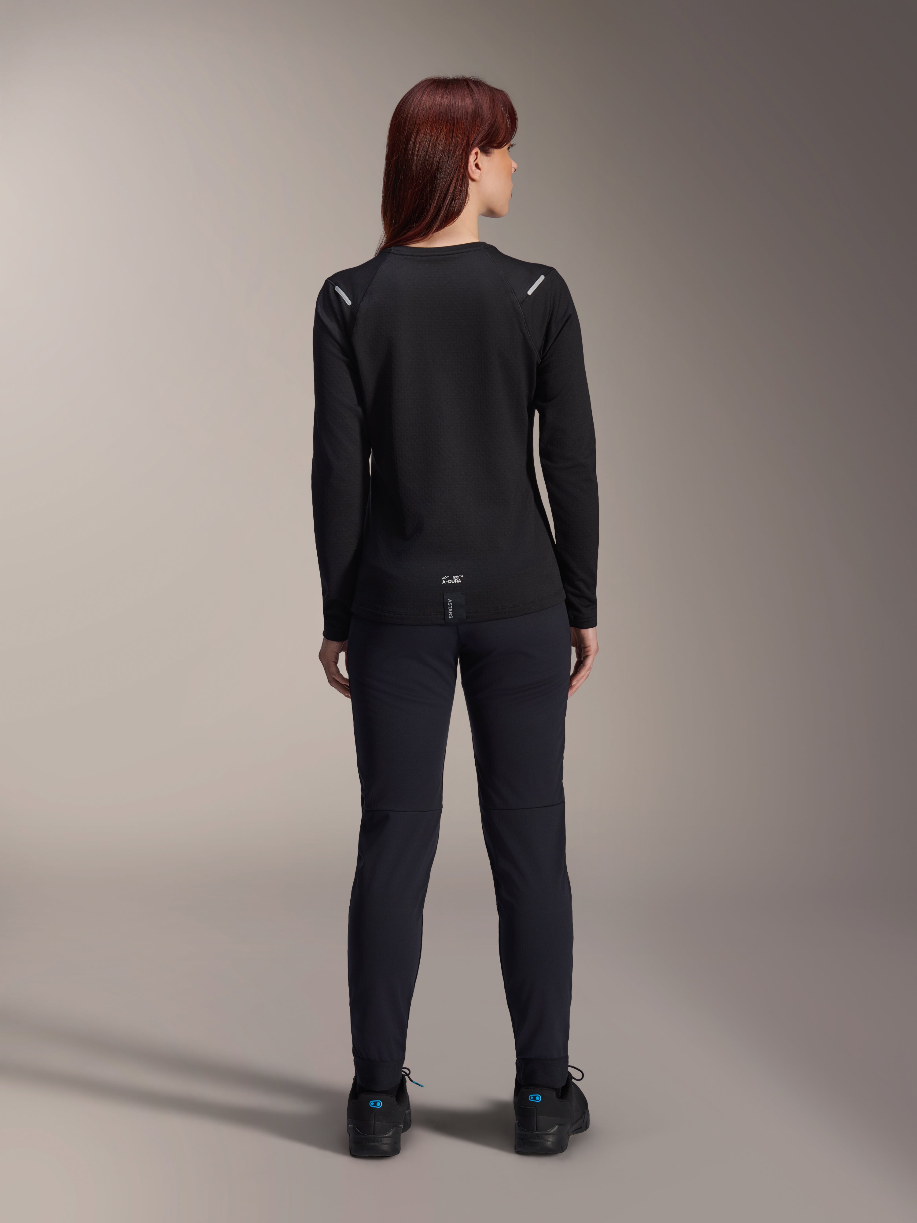 Alpinestars Damen Stella A-Dura Thermo-Trikot, Rückansicht, Schwarz, langärmliges Radtrikot mit isoliertem Fleece, getragen mit passender schwarzer Hose und Schuhen, mit kleinem A-Dura-Branding am unteren Rücken und reflektierenden Akzenten an den Schultern, speziell für Mountainbike-Fahrten von Frauen entwickelt.