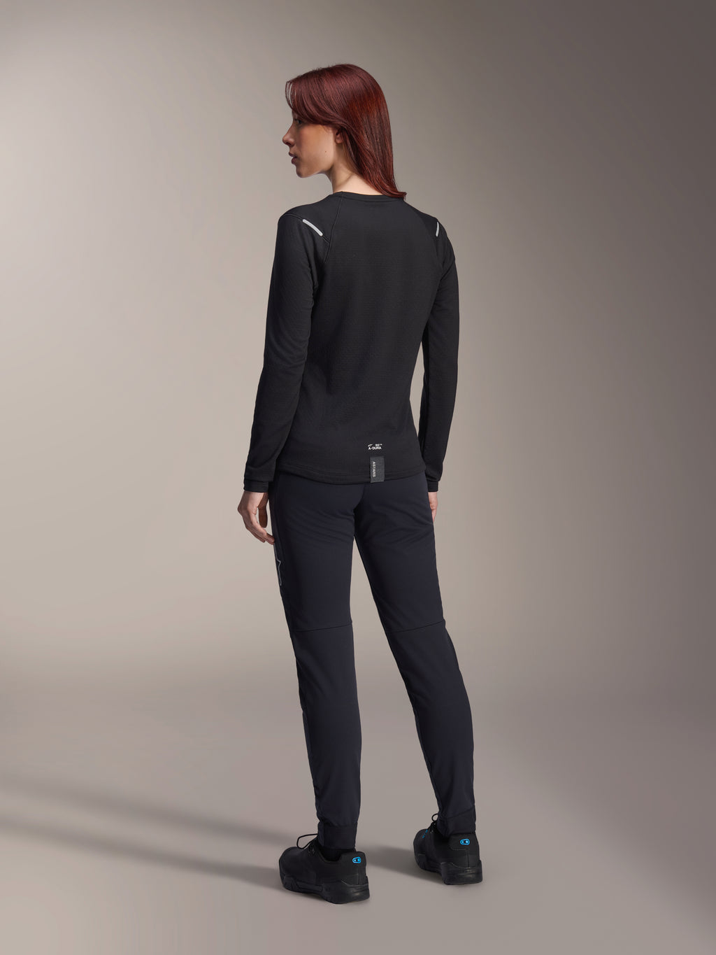 Alpinestars Damen Stella A-Dura Thermo-Trikot, langärmliges MTB-Trikot in Schwarz, Rückansicht mit reflektierenden Schulterakzenten und verlängertem Rückenteil, getragen mit schwarzer Hose und Schuhen, Performance-Ausrüstung fürs Mountainbiking