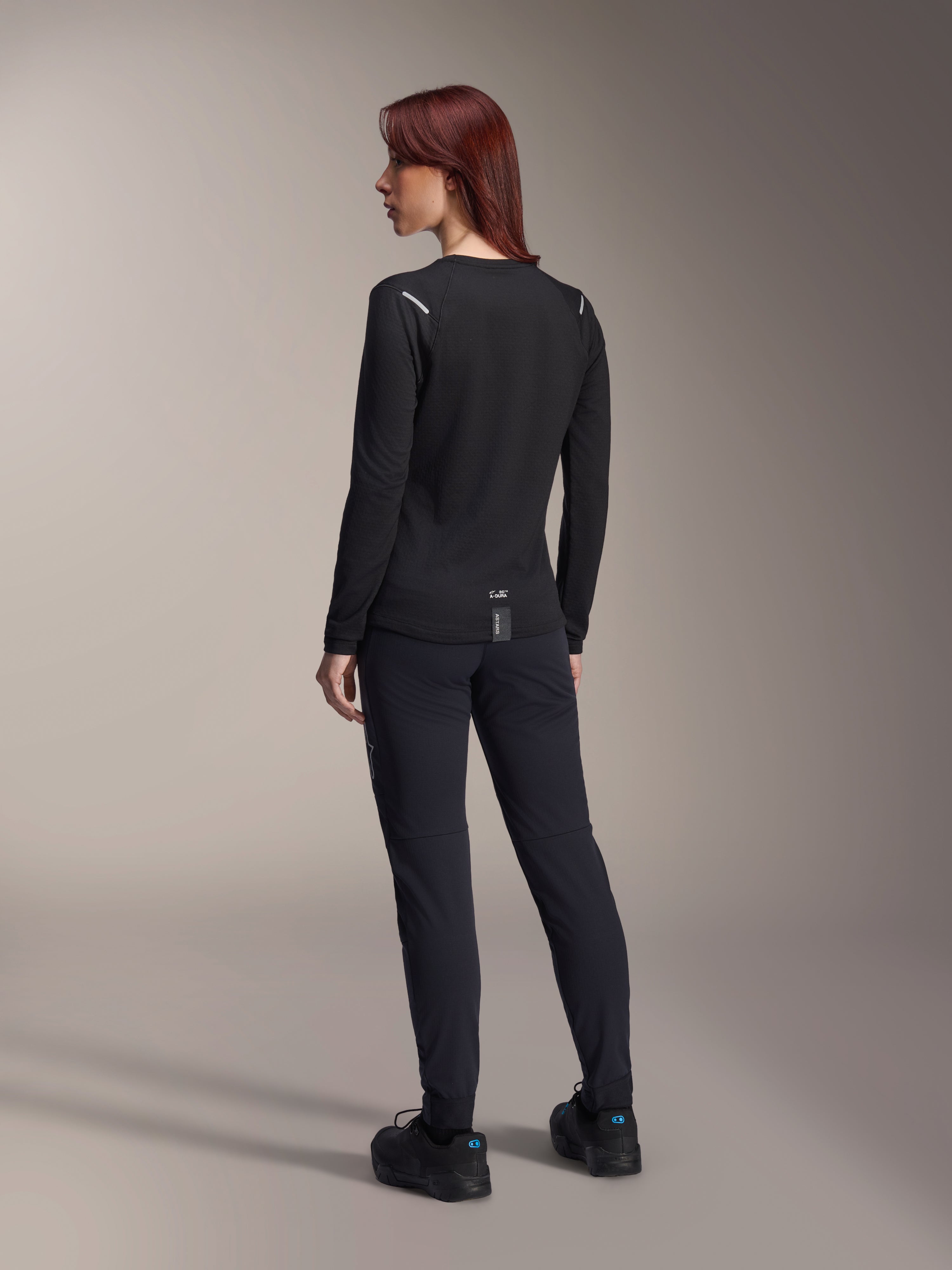 Alpinestars Damen Stella A-Dura Thermo-Trikot, langärmliges MTB-Trikot in Schwarz, Rückansicht mit reflektierenden Schulterakzenten und verlängertem Rückenteil, getragen mit schwarzer Hose und Schuhen, Performance-Ausrüstung fürs Mountainbiking