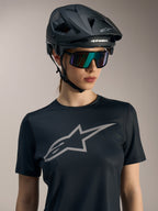 Schwarzes Stella A-Dura Ride Damen-Kurzarmtrikot mit großem grauem Brustlogo, dazu MTB-Helm und Sonnenbrille.