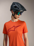 Oranges Alpinestars Stella A-Dura Ride Kurzarmtrikot mit rotem Logo, schwarzem MTB-Helm und schillernder Sonnenbrille.