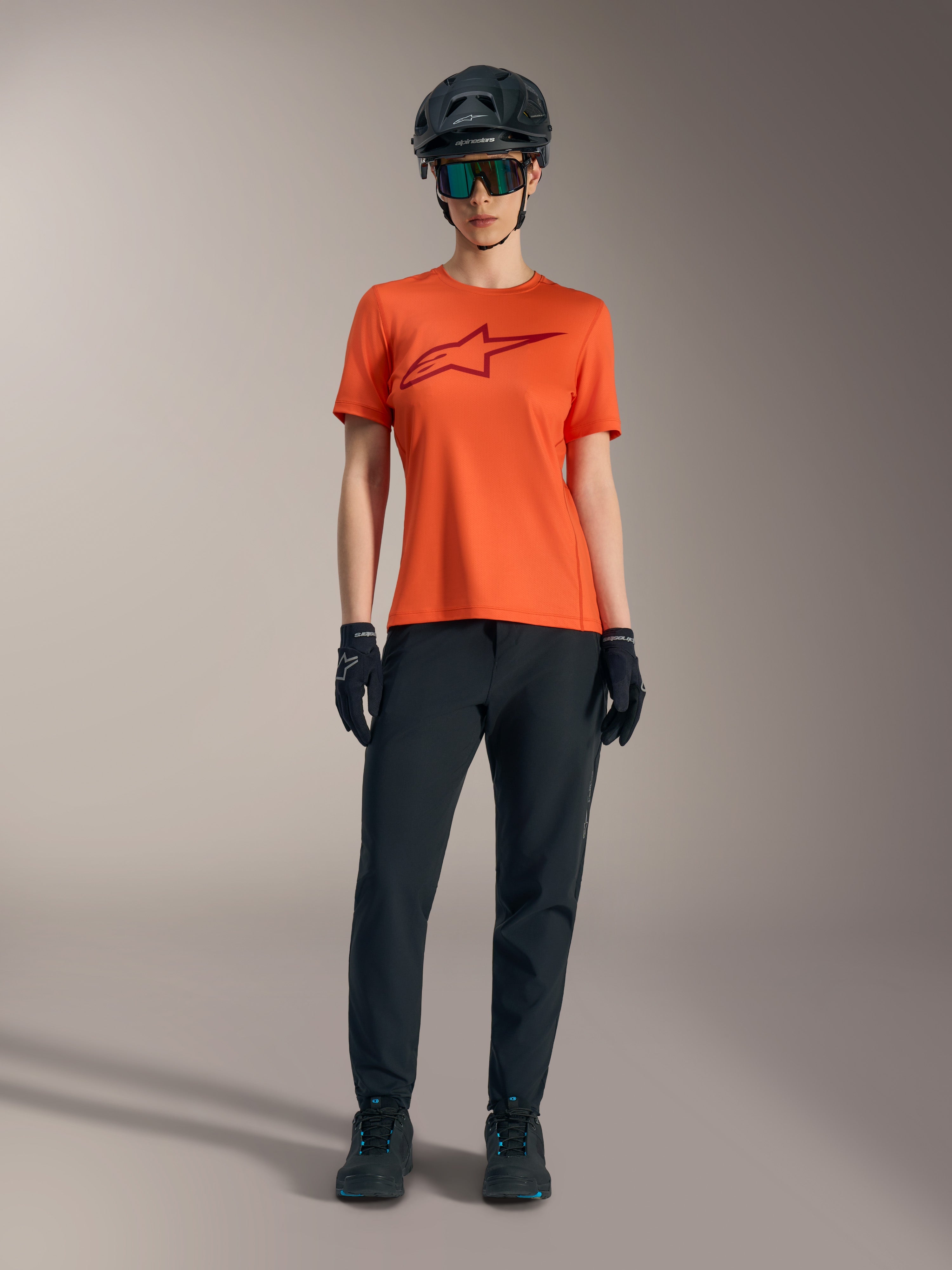Oranges Stella A-Dura Ride Kurzarmtrikot mit weinrotem Logo, dazu schwarze MTB-Ausrüstung, Helm und Handschuhe.
