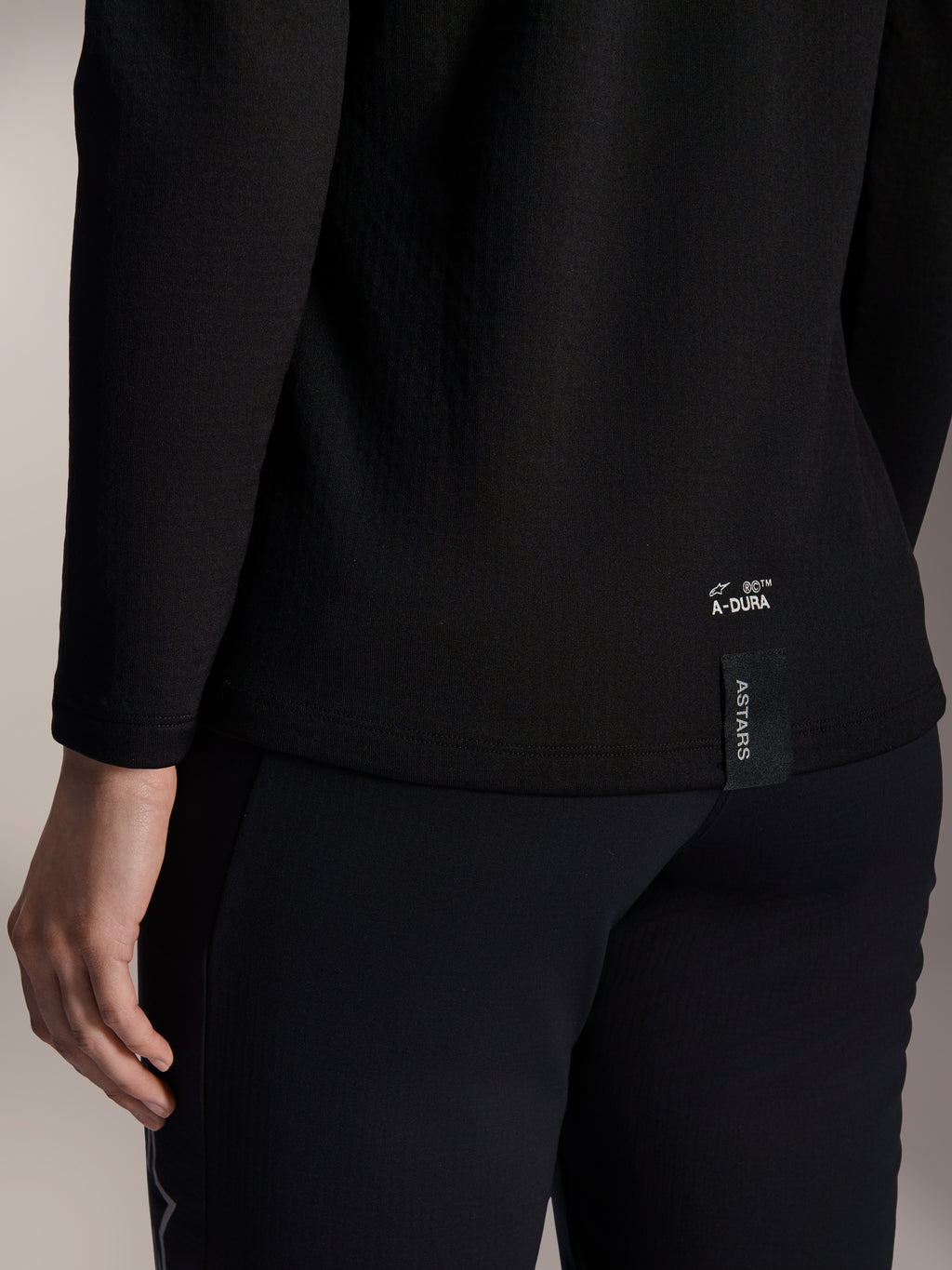 Alpinestars Stella A-Dura Thermal Jersey für Damen, schwarzes MTB-Thermo-Trikot, Nahaufnahme des unteren Rückensaums mit weißem A-DURA-Branding und vertikalem ASTARS-Logo-Tag, entwickelt für Mountainbiking bei kaltem Wetter
