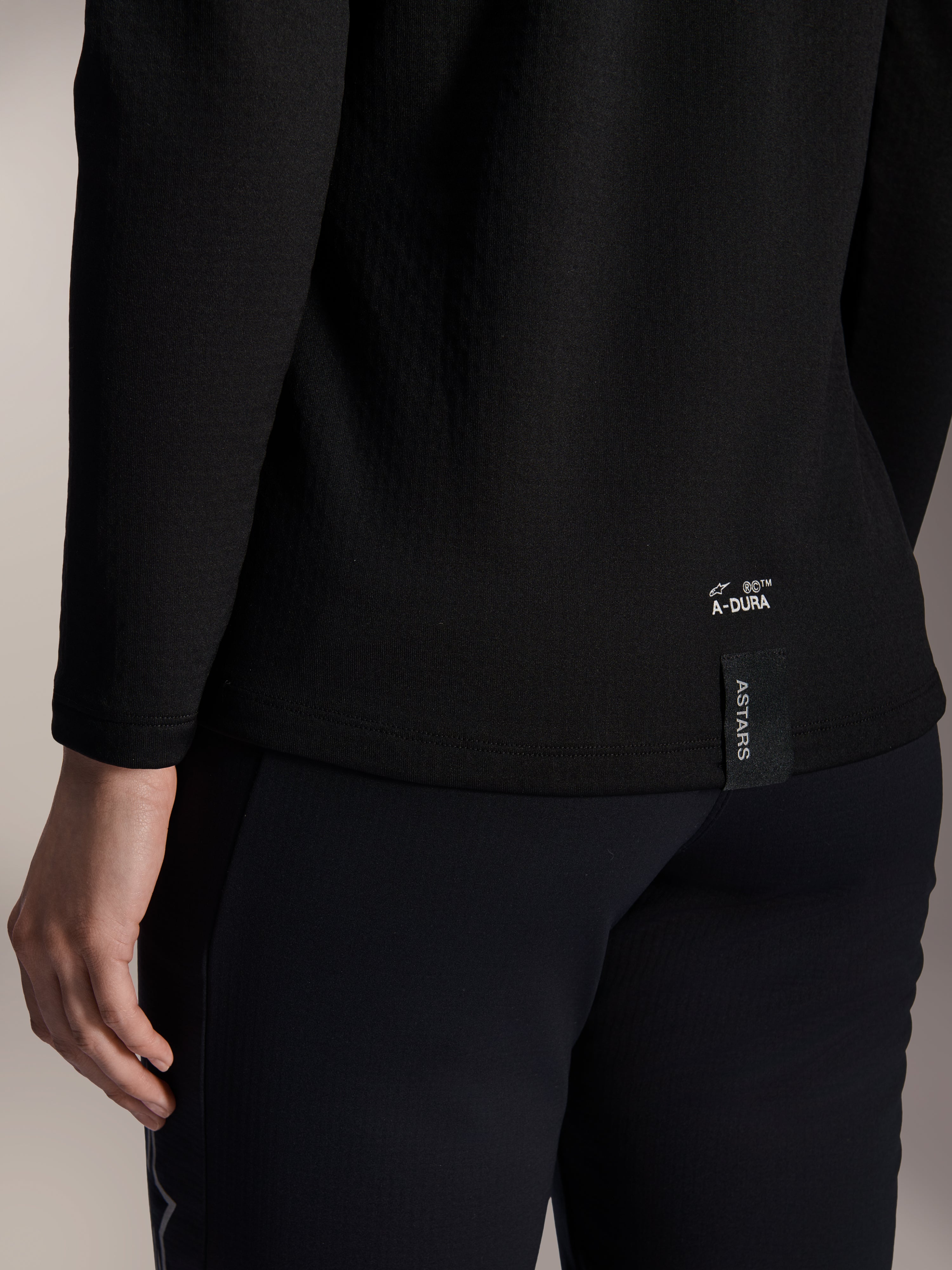 Alpinestars Stella A-Dura Thermal Jersey für Damen, schwarzes MTB-Thermo-Trikot, Nahaufnahme des unteren Rückensaums mit weißem A-DURA-Branding und vertikalem ASTARS-Logo-Tag, entwickelt für Mountainbiking bei kaltem Wetter
