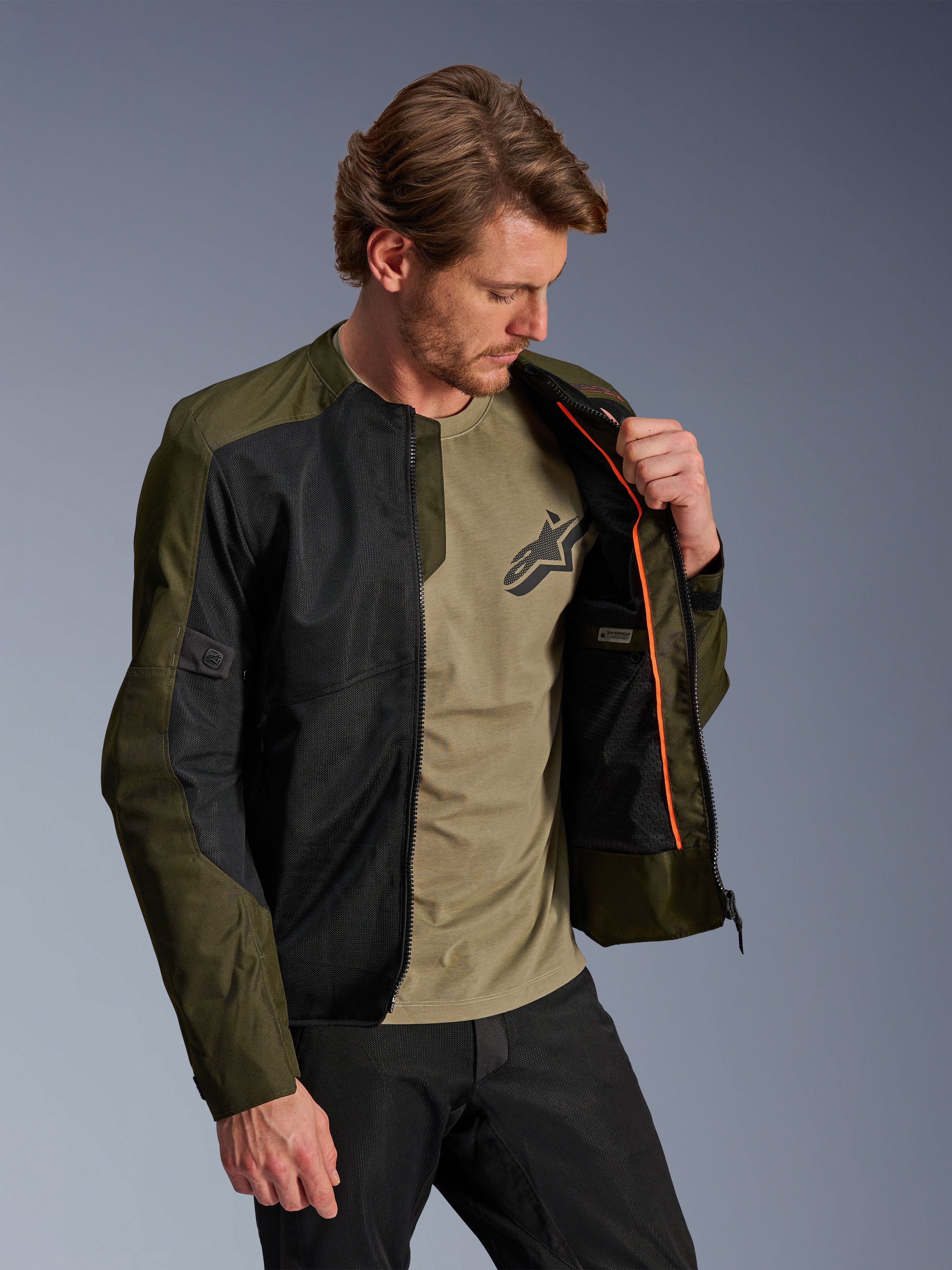 Alpinestars C-1 Air Jacke in Khaki und Schwarz, getragen von einem männlichen Model zur Darstellung des orangefarbenen Innenfutters und des wasserdichten Fachs, mit großen Tech-Mesh-Einsätzen für Luftzirkulation, urbaner zeitgenössischer Motorradstil kombiniert mit einem passenden olivfarbenen T-Shirt und einer schwarzen Hose