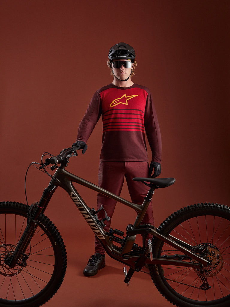 Bordeauxfarbenes A-Dura Elite 2.0 Langarm-MTB-Trikot mit rotem Brustpanel und gelbem Logo, abgebildet mit Mountainbike.