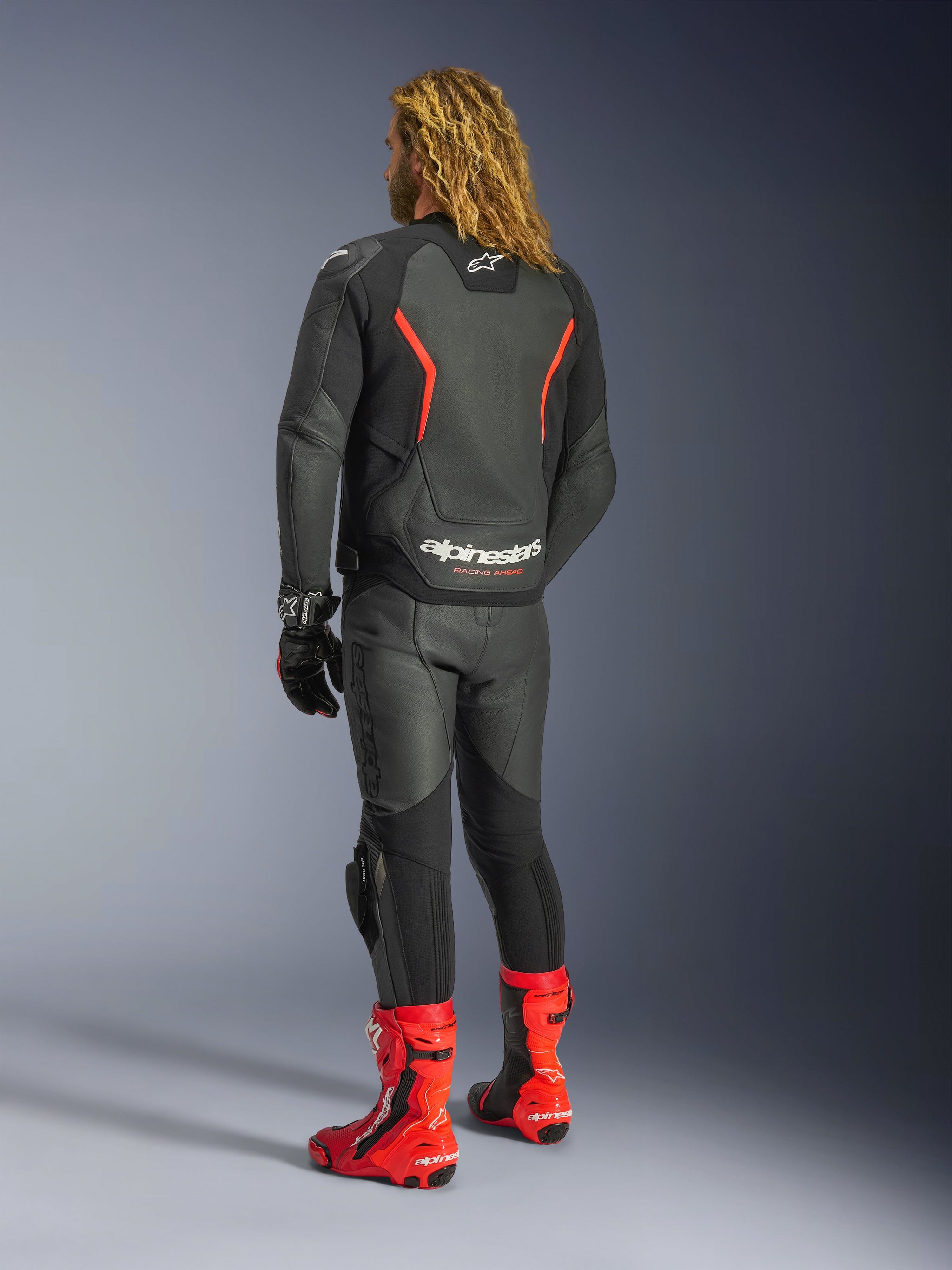 Alpinestars GP Force V2 Lederjacke, Schwarz-Rot-Fluo, Rückansicht getragen von einem Fahrer mit passender schwarzer Lederhose, roten Rennstiefeln und schwarzen Handschuhen, mit markanten roten Akzenten und Alpinestars-Branding auf dem Rücken, Motorradausrüstung in Sport-Passform