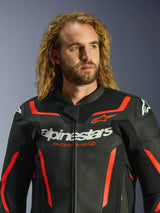 Alpinestars GP Force V2 Lederjacke in Schwarz und Rot Fluo, getragen von einem männlichen Model mit langen lockigen blonden Haaren, markantes weißes Alpinestars-Branding und „RACING AHEAD“-Schriftzug auf der Brust, hochwertige Motorrad-Lederjacke für sportliche Performance.
