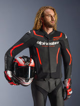Alpinestars GP Force V2 Lederjacke, Schwarz und Rot Fluo, getragen von einem männlichen Model, das einen passenden Helm hält, mit weißen Alpinestars-Logos und roten Akzenten, kombiniert mit einer Lederhose und Rennhandschuhen, Sport-Motorradausrüstung für Straße und Rennstrecke.