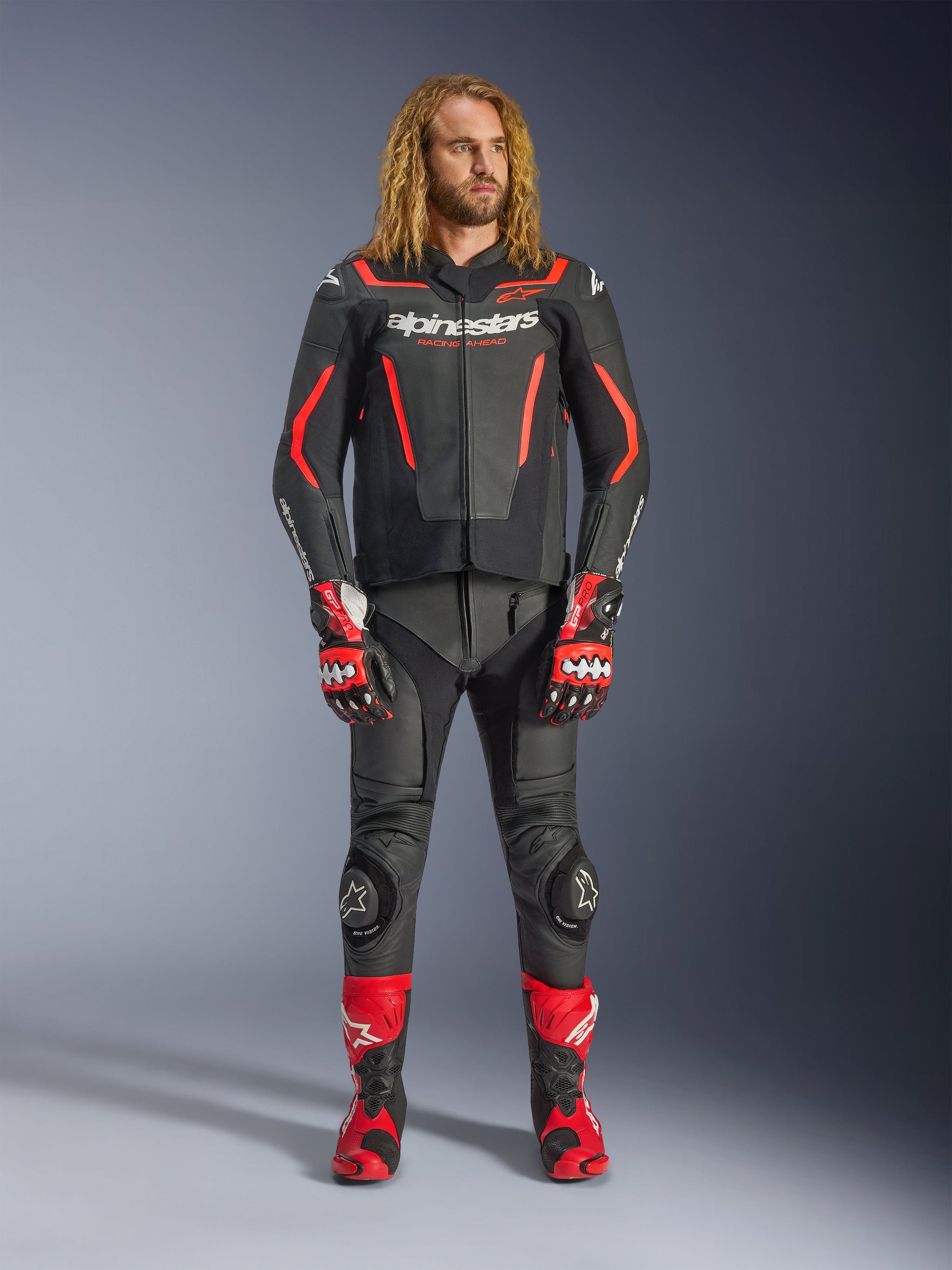 Männliches Model in der Alpinestars GP Force V2 Lederjacke in Schwarz und Rot Fluo, kombiniert mit passender Lederhose, roten Rennhandschuhen und roten Performance-Stiefeln, professionelle Motorrad-Sportbekleidung