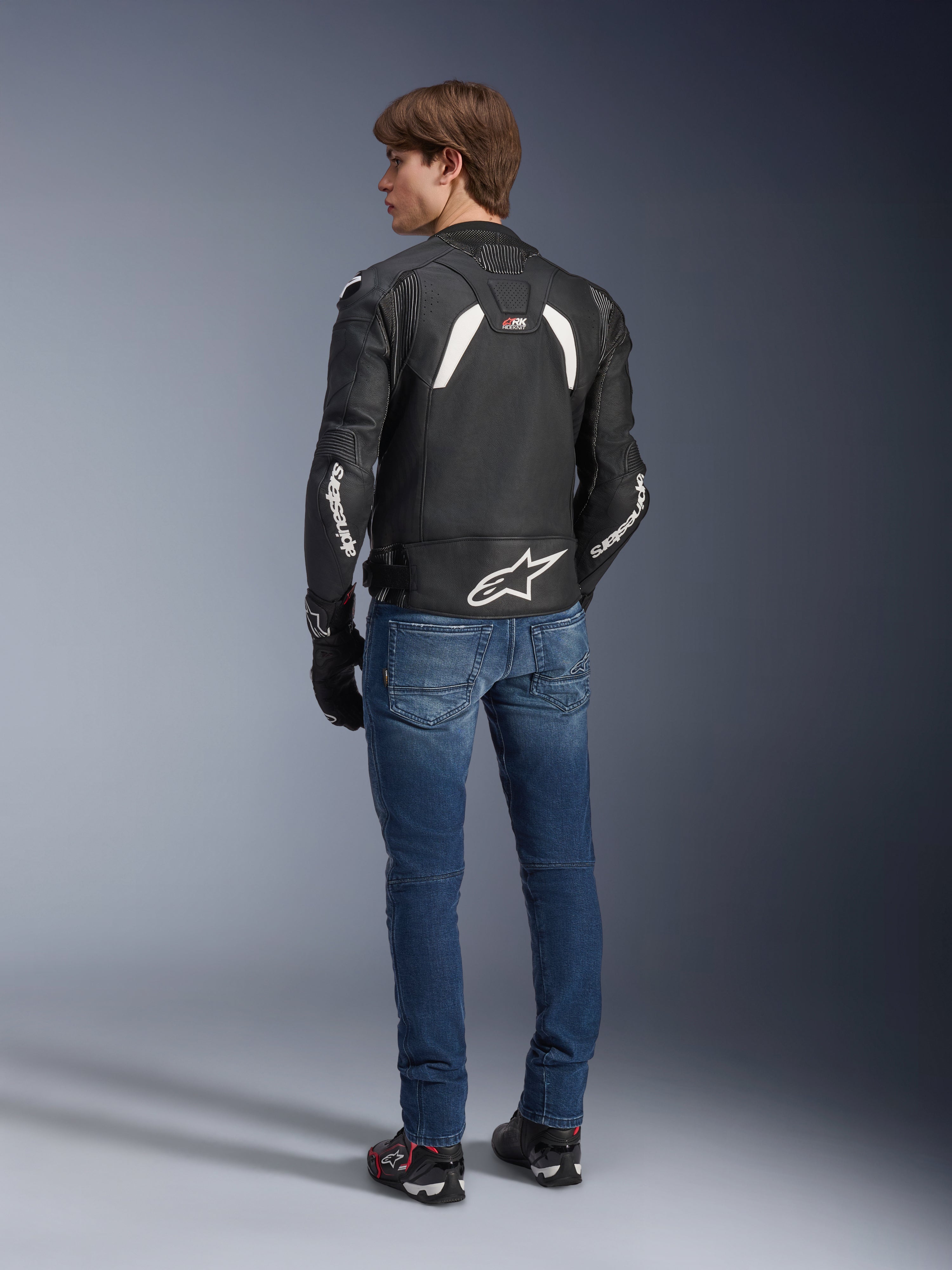 Alpinestars GP Plus R V4 Rideknit Leather Jacket in Black and White, rear view on a model, featuring white Alpinestars branding on sleeves and a large star logo on the lower back, worn with blue jeans and black riding shoes, technical motorcycle gear.