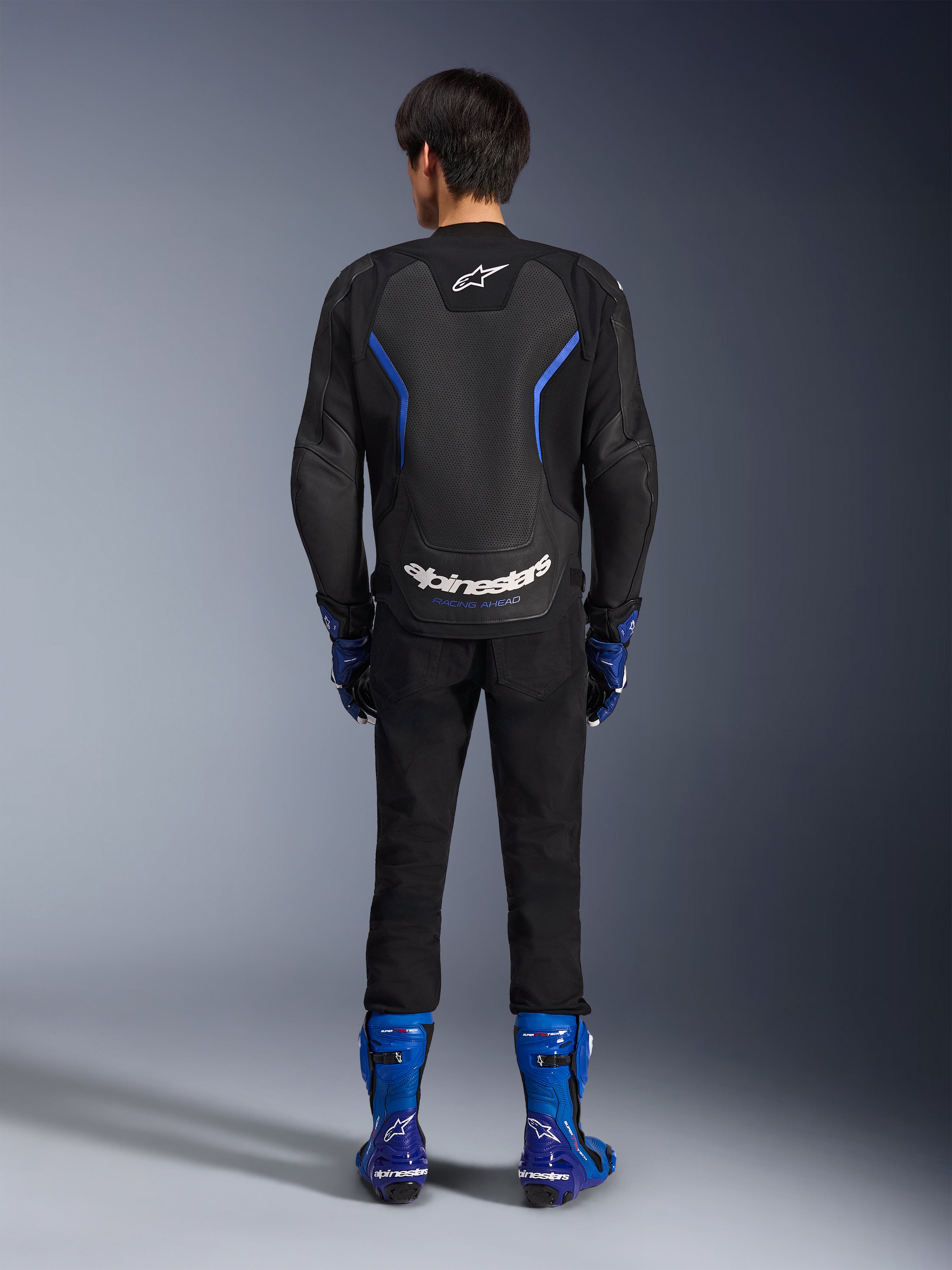 Alpinestars GP Force V2 Airflow Leather Jacket, Black Metallic Blue, rear view showing perforated leather panels and white branding, worn with blue racing boots and black pants, sport motorcycle racing gear