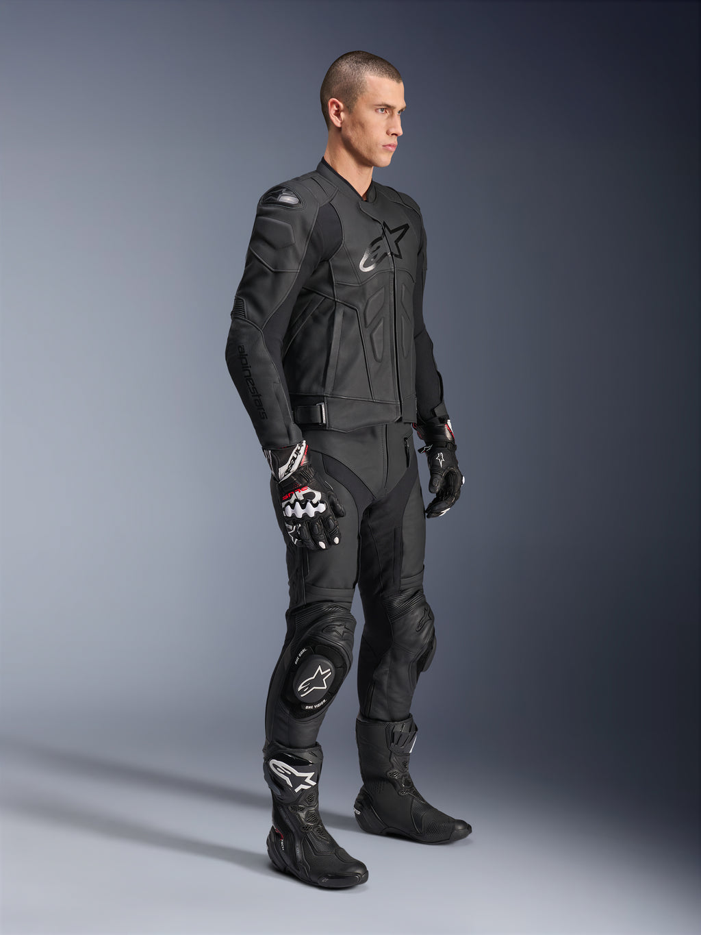 Alpinestars Dusk Lederjacke, Schwarz, getragen von einem männlichen Model mit passender Motorrad-Lederhose, Rennhandschuhen und Stiefeln, markantes Stern-Logo auf der Brust, ergonomisches Sport-Design für die Straße